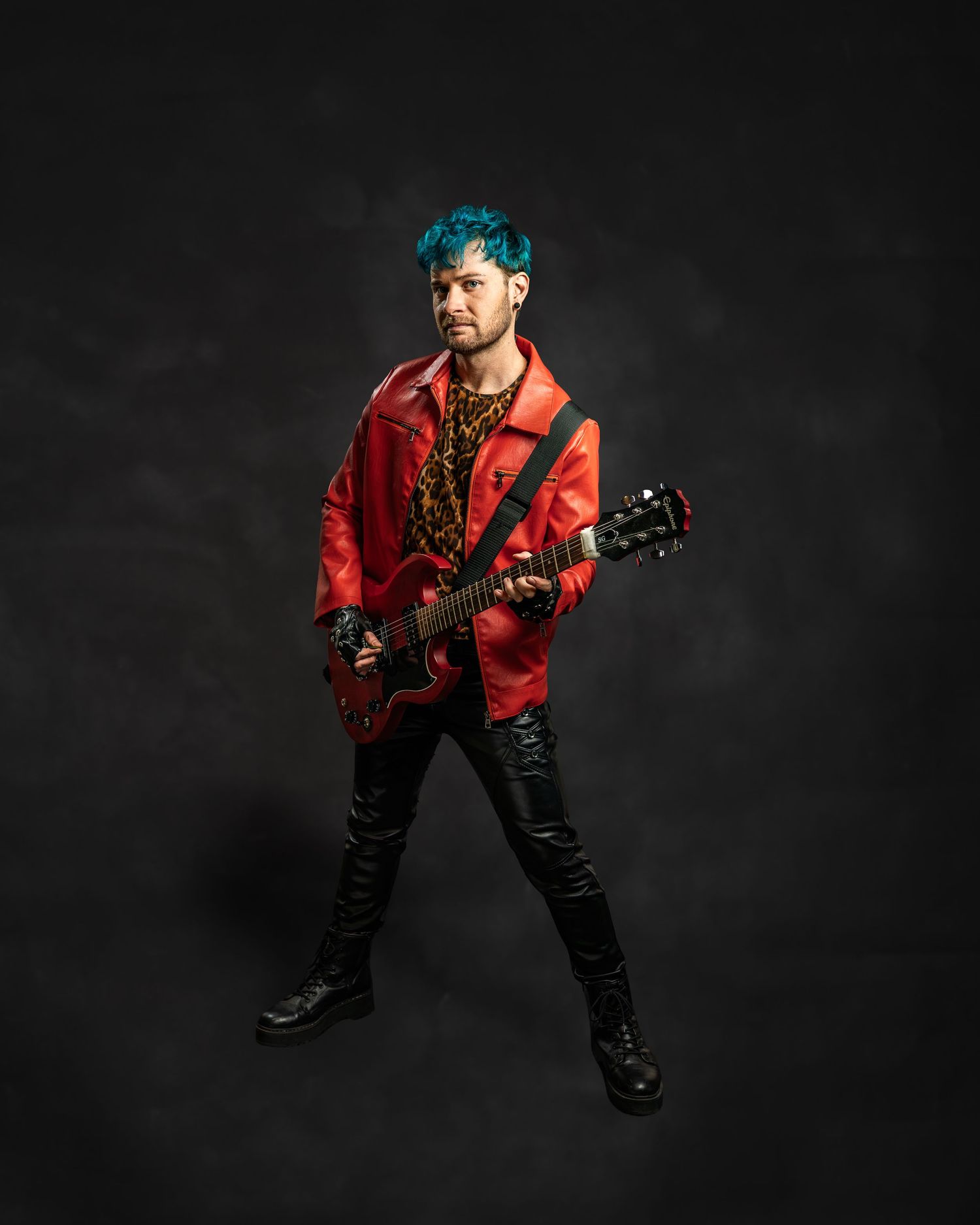 Musician with teal hair in red leather jacket plays red electric guitar against dark studio background.