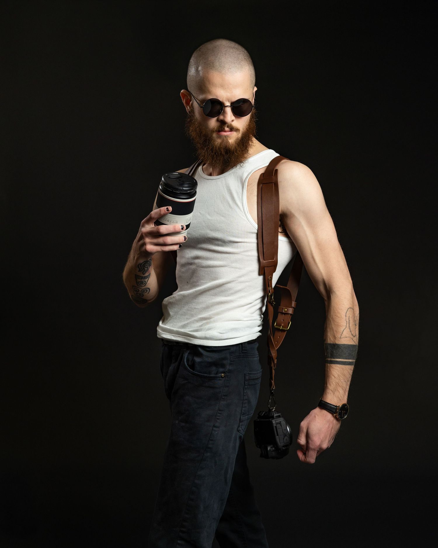 Muscular photographer in white tank top and sunglasses poses with camera against dark background.