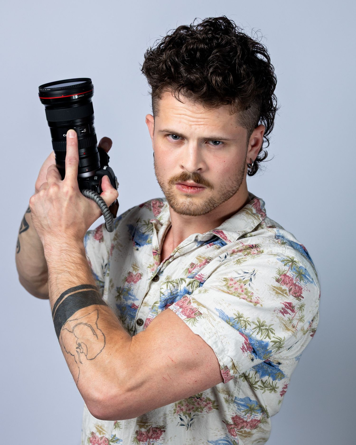 Tattooed man in floral shirt aiming a camera lens forward, curly hair, studio photography portrait.