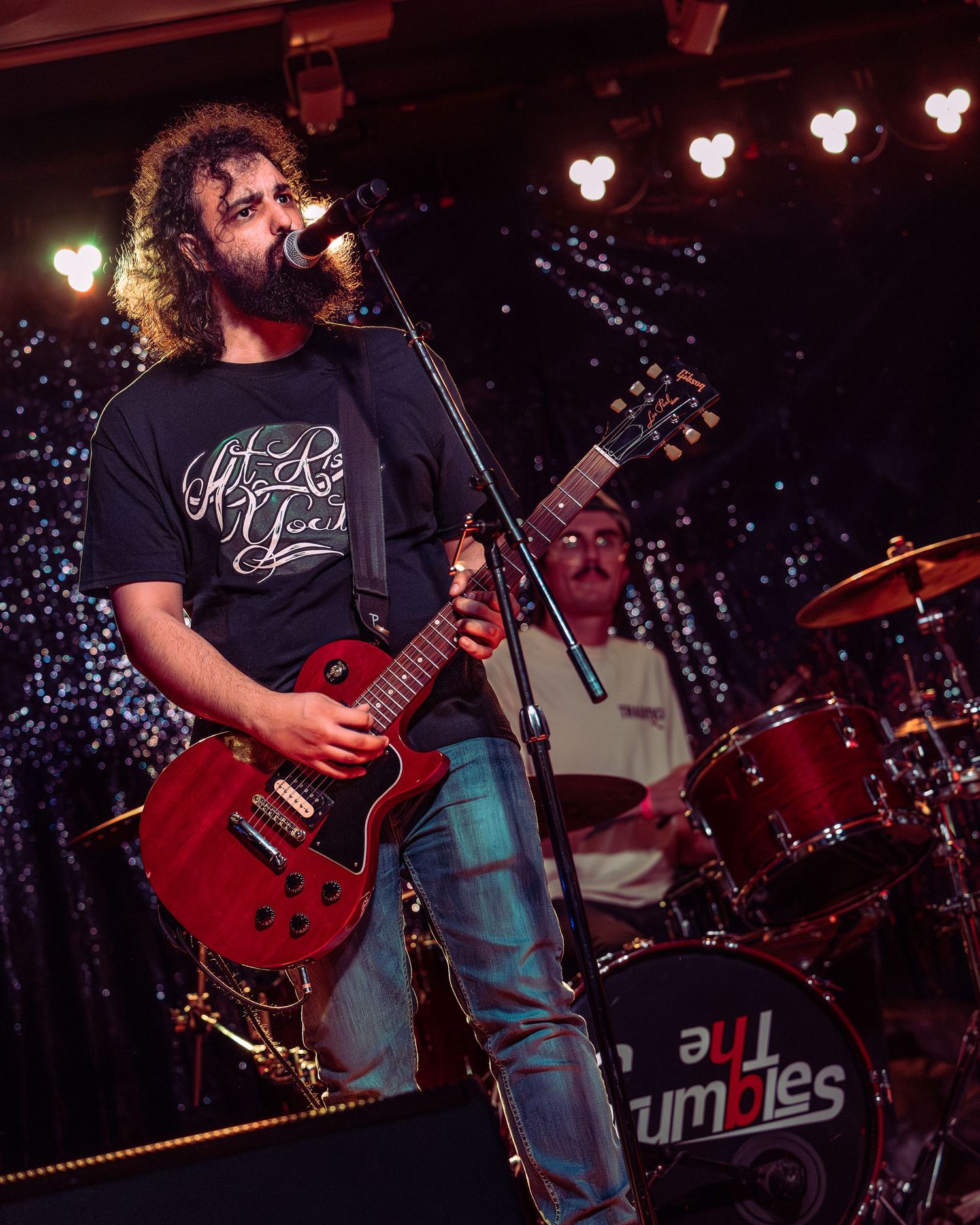 Curly-haired guitarist singing into microphone on stage playing red electric guitar with drummer visible behind.