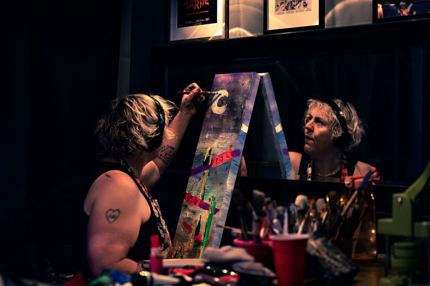 Artist painting a colorful canvas on an easel in a dimly lit blue room with tattoos visible.
