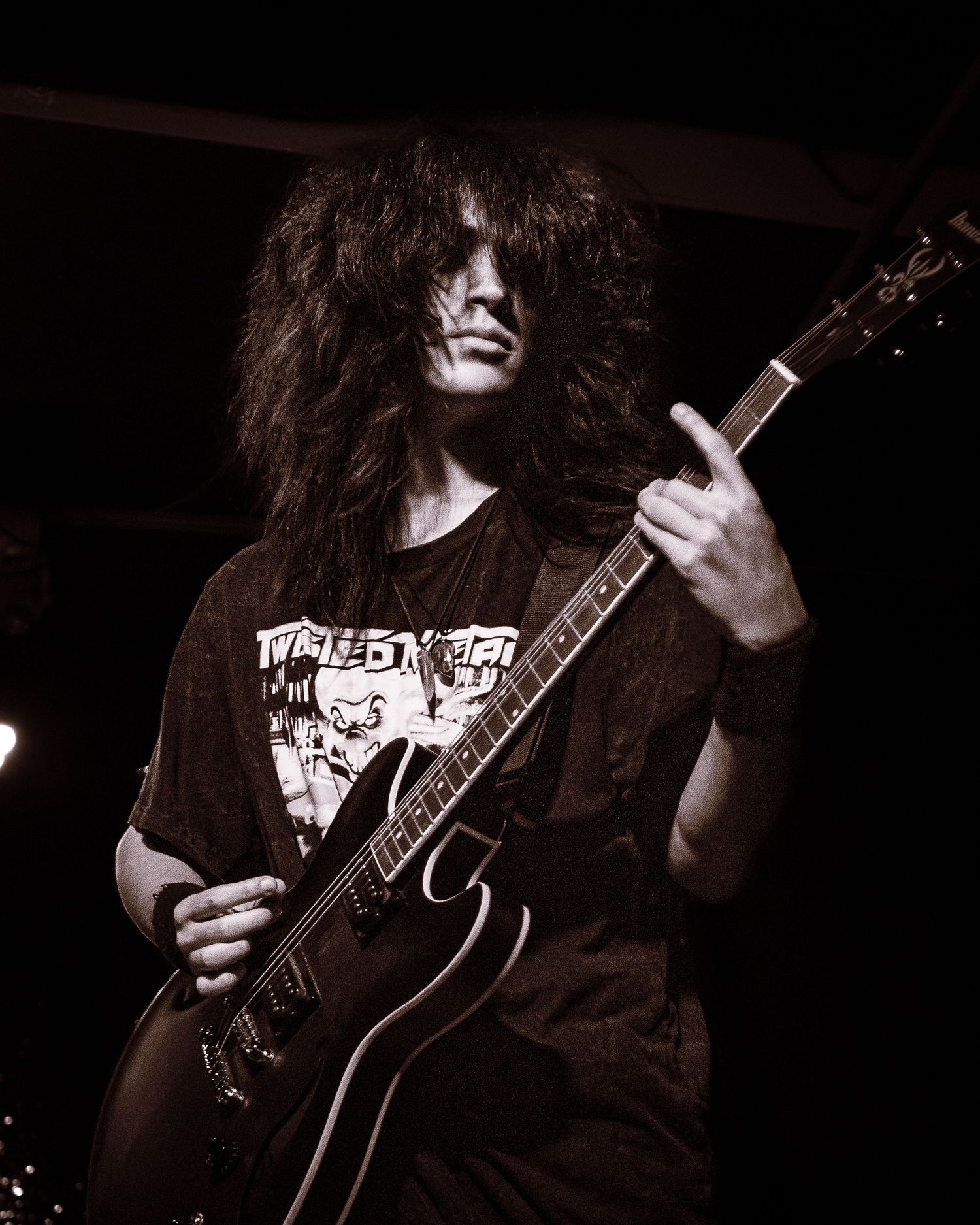 A long-haired guitarist plays electric guitar on stage in a black band t-shirt, captured in moody sepia tones.