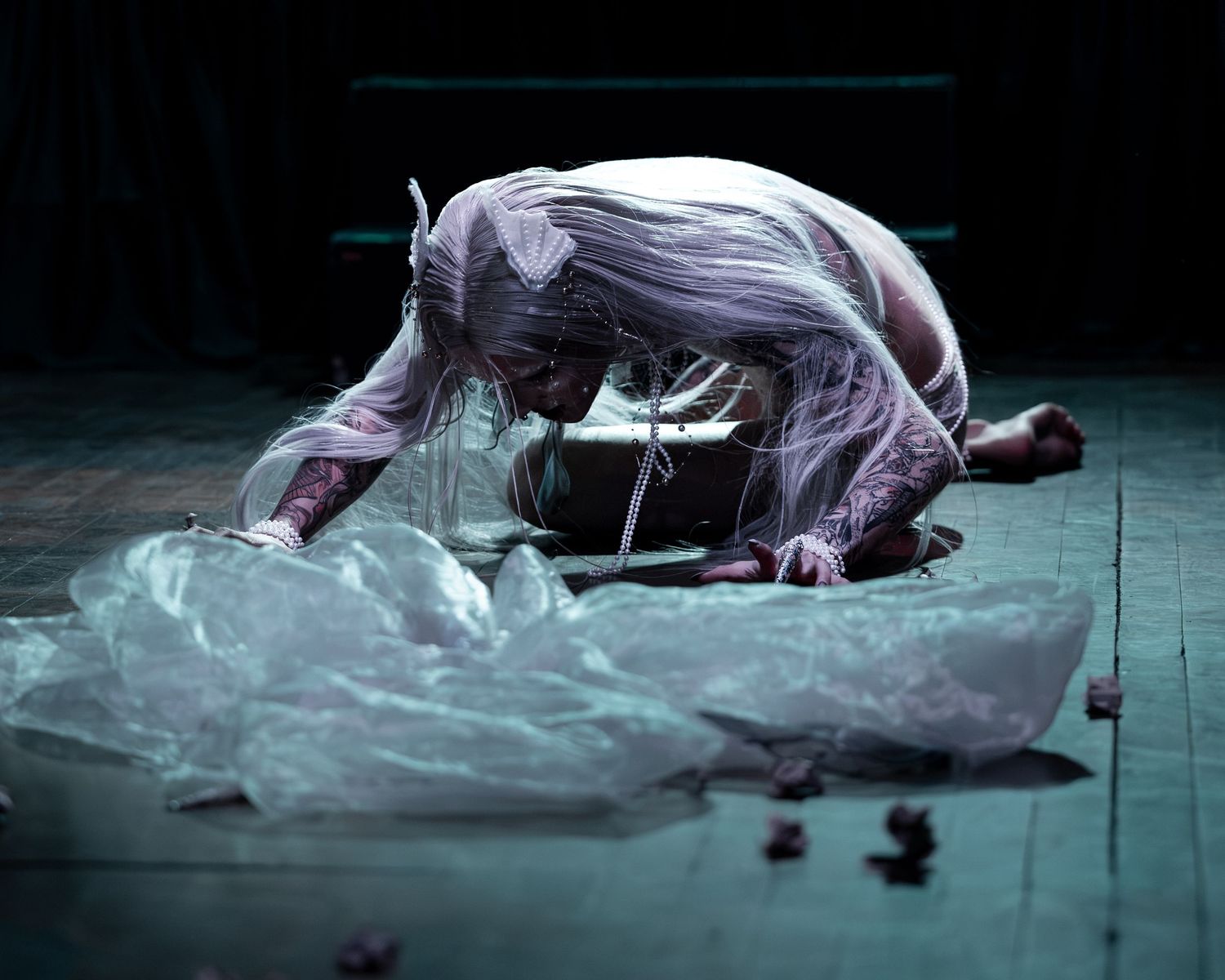 Dramatic theater performer in white translucent costume crouched on dark stage floor amid flowing fabric.