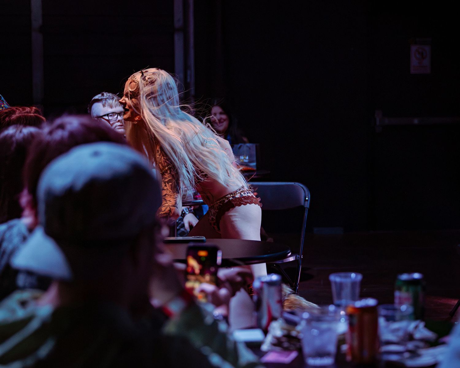 Blonde tattooed performer interacting with audience at dimly lit cabaret show table setting.