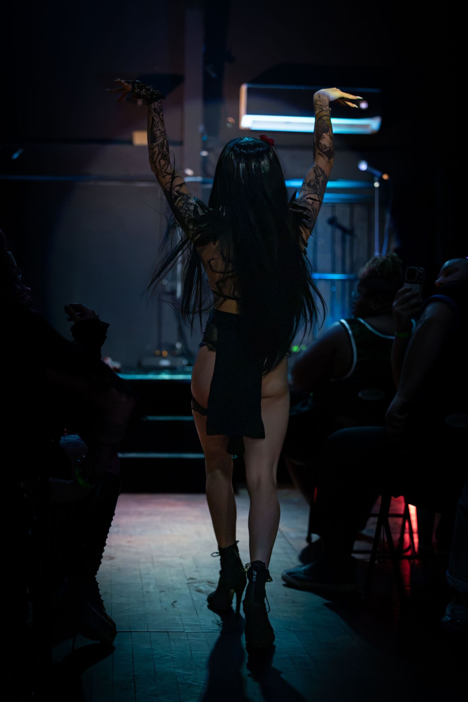 Gothic performer with tattooed arms raised stands on dimly lit stage before a seated audience.