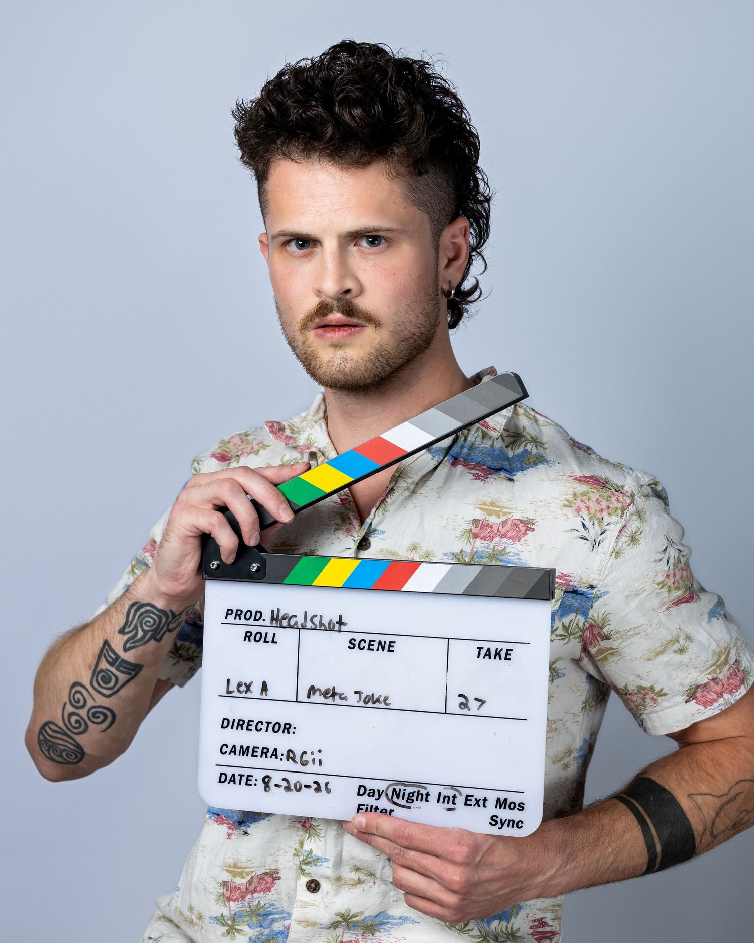 Man with tattoos and mustache holds a film clapperboard, wearing a floral shirt in a studio setting.