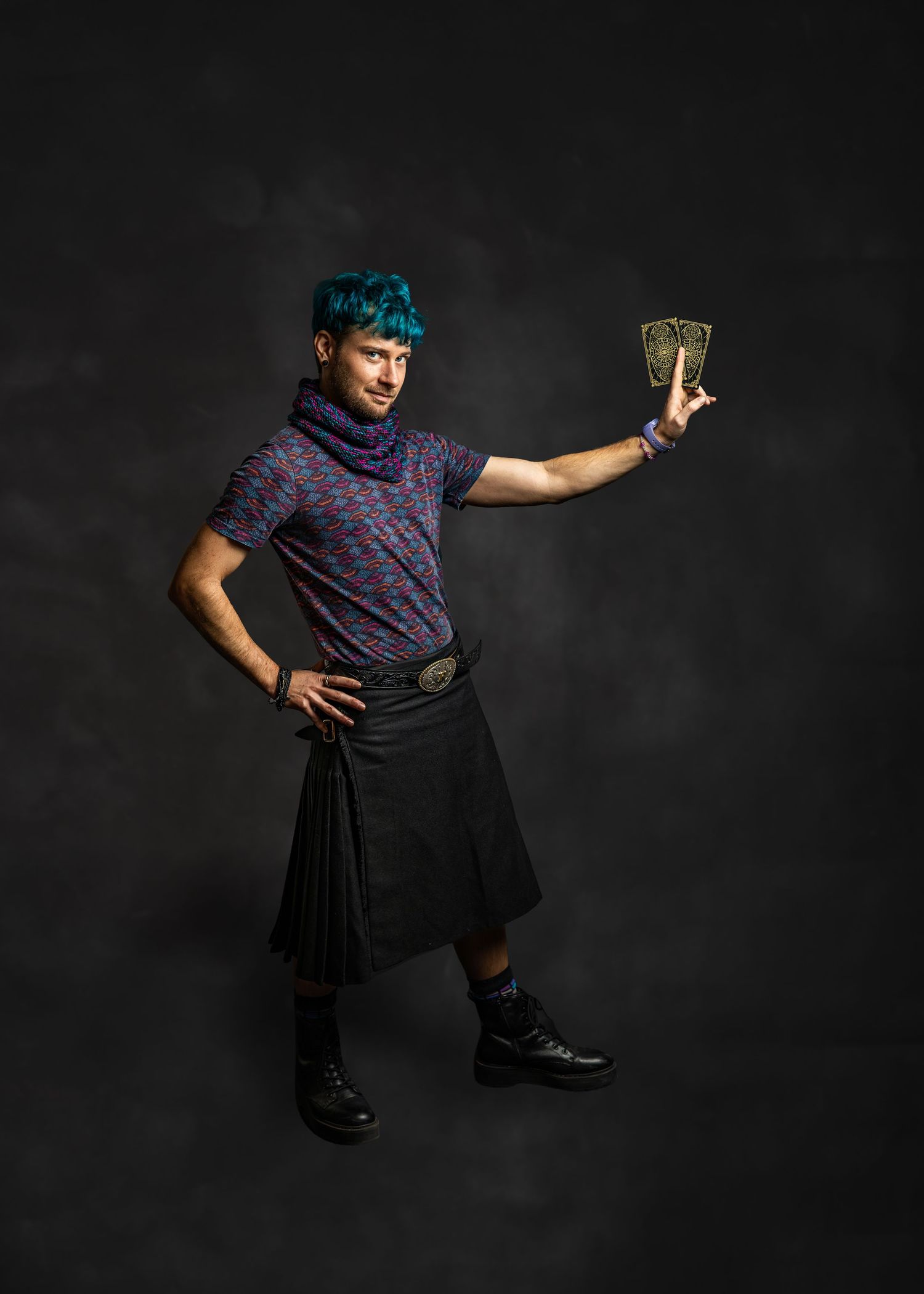 Person with teal hair wearing a black kilt and patterned shirt, holding a decorative card while posing dramatically.