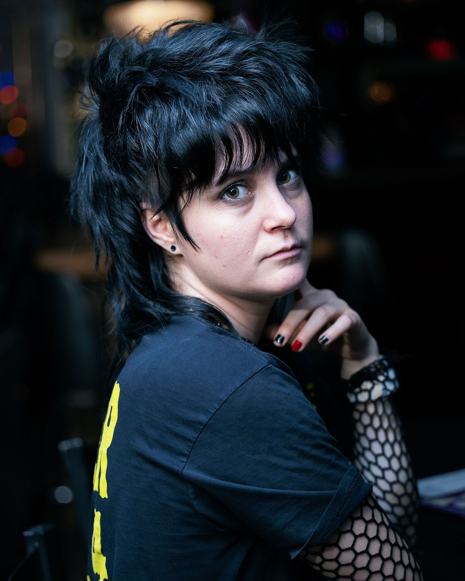 Young woman with black mullet hair, fishnet sleeves, and red nails poses in a dark venue wearing a black graphic tee.