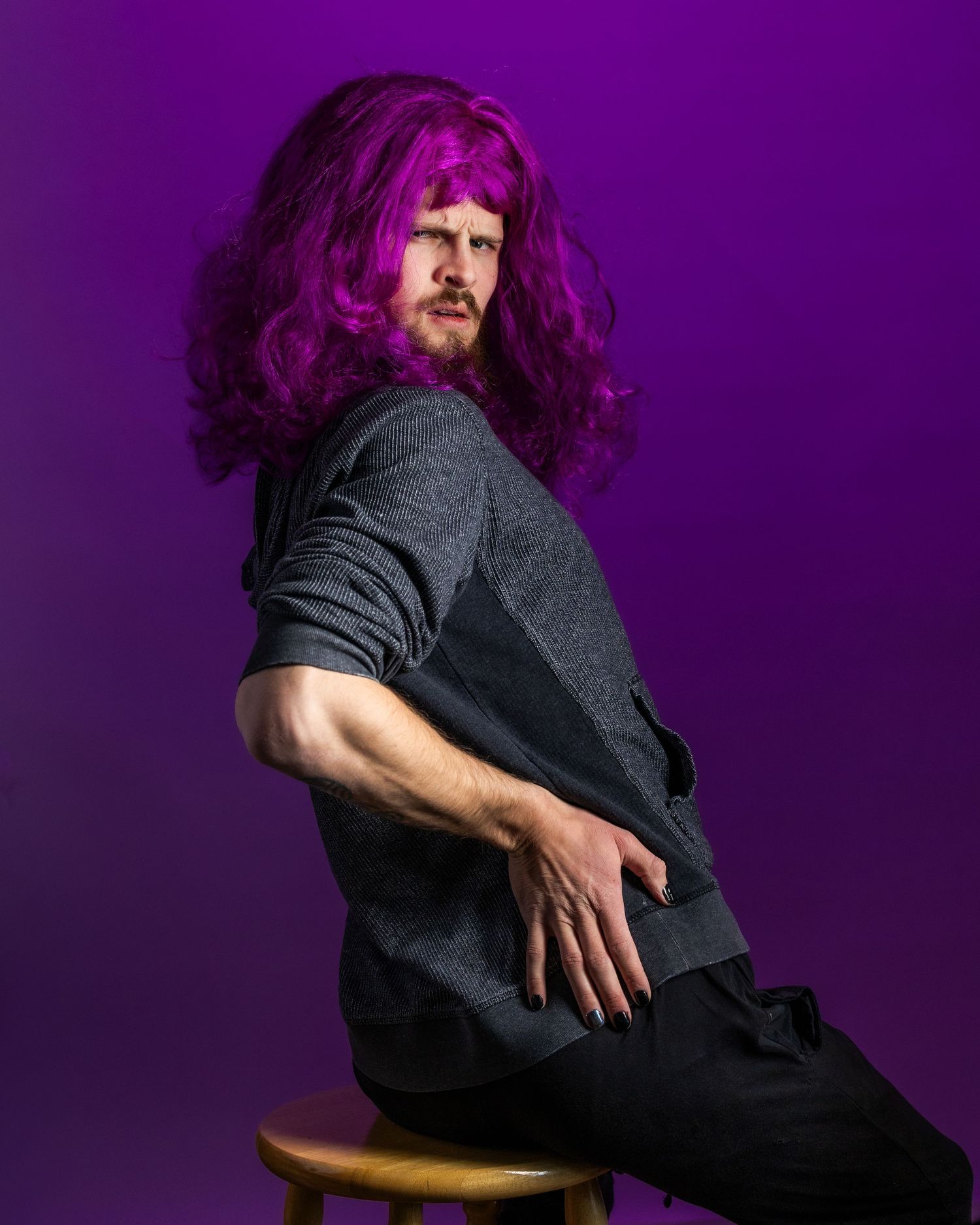 Artistic portrait against purple backdrop with vibrant magenta hair and dark clothing in contemplative pose.