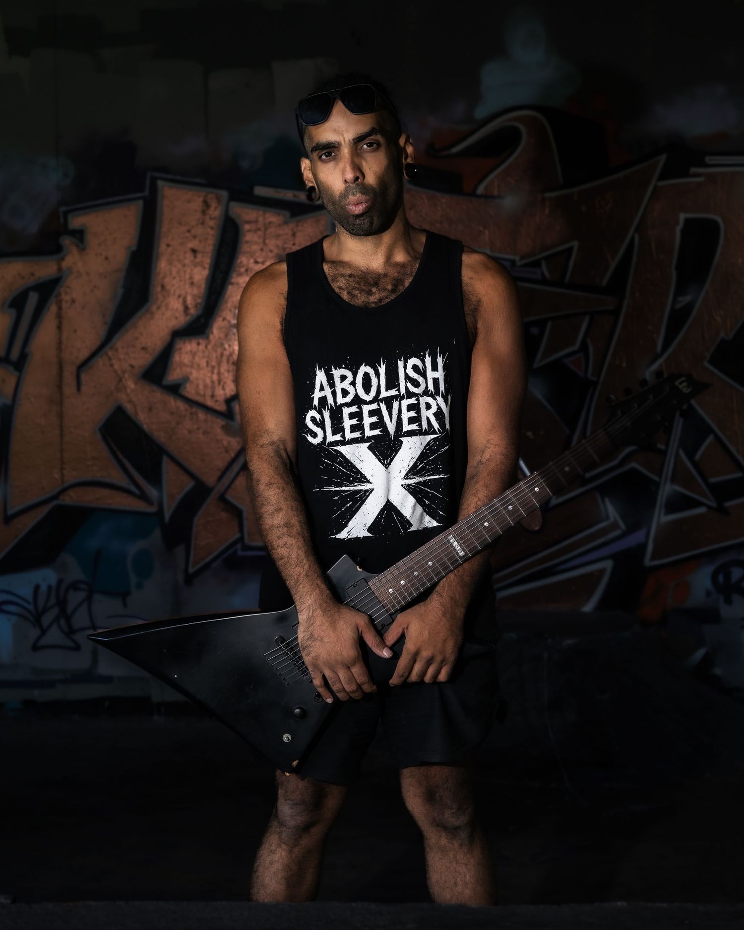 A person in a dark room wearing a sleeveless shirt with tattoos visible on their arms.