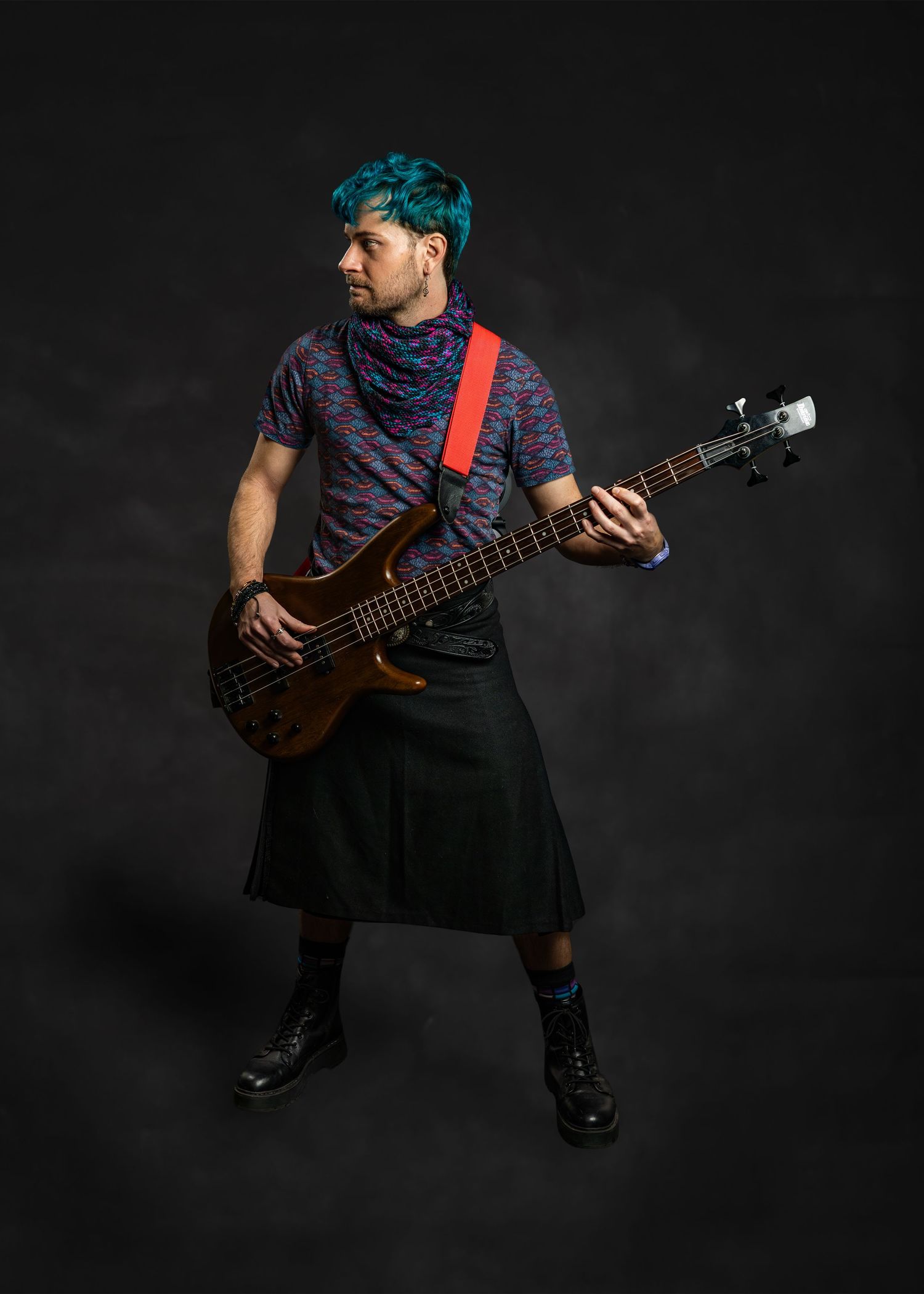 Teal-haired male bassist in black kilt with red guitar strap posing in dark studio.