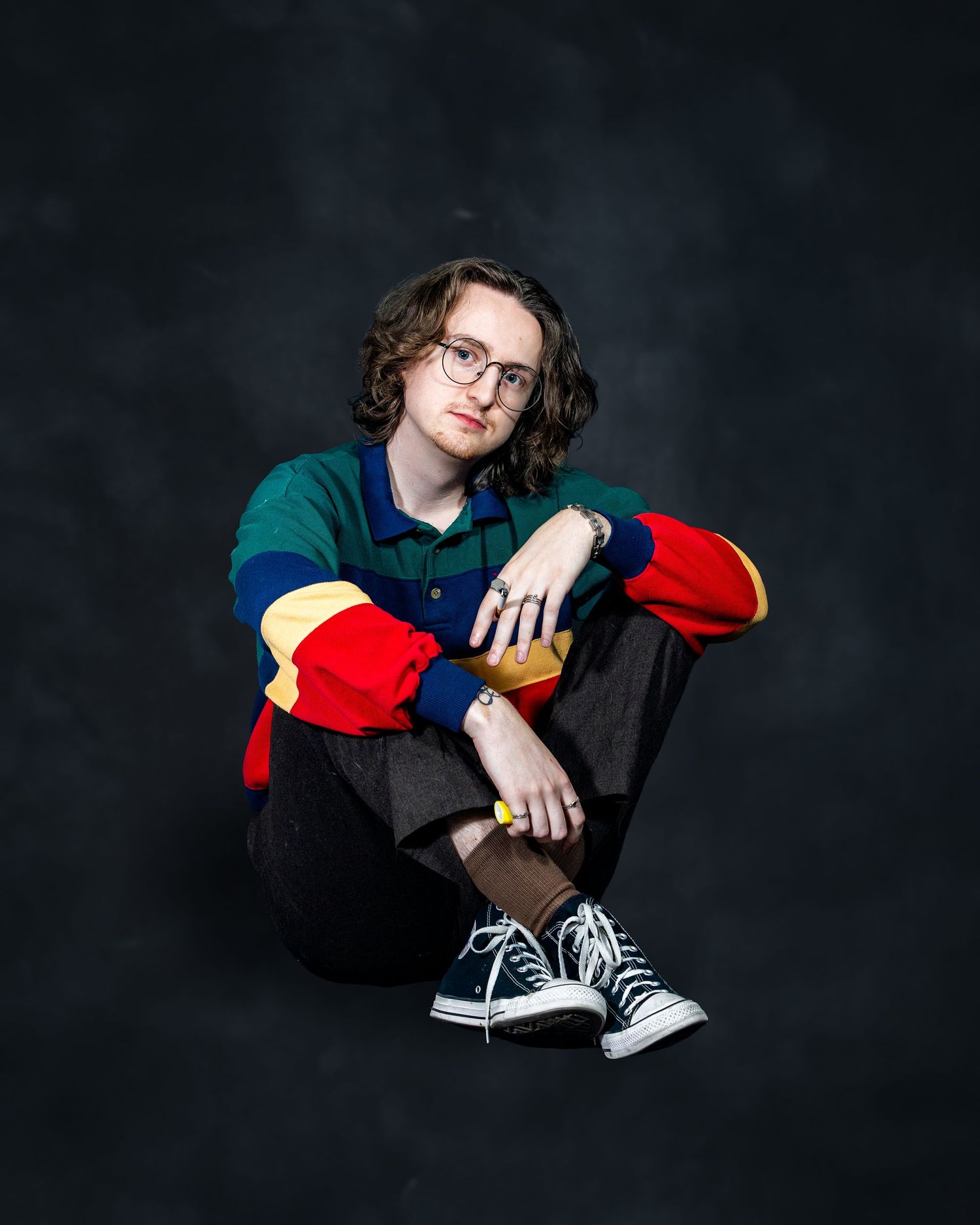 Young person in colorful retro polo shirt sitting cross-legged in studio portrait.