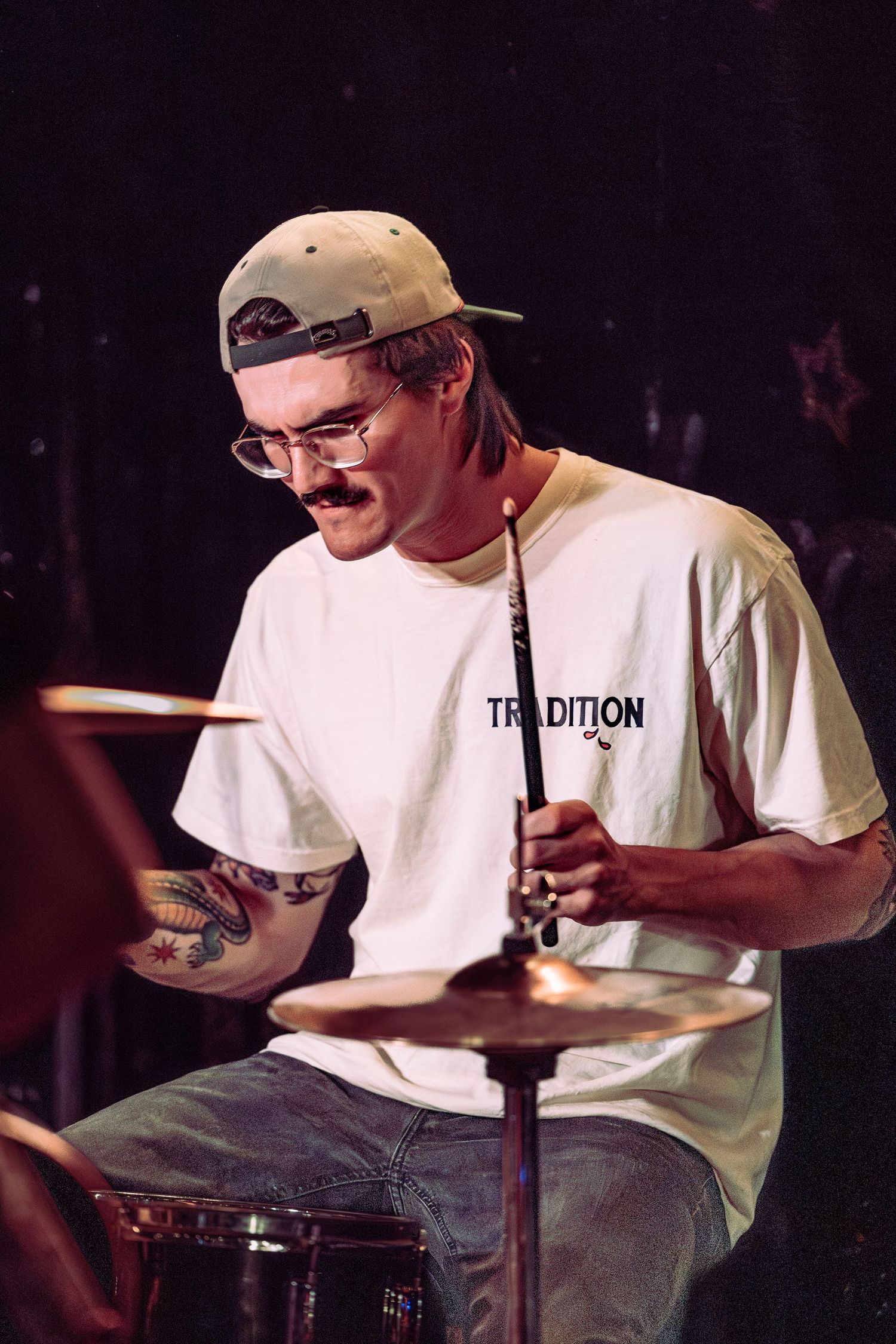 Drummer with mustache and cap plays cymbals on stage wearing white Tradition t-shirt.