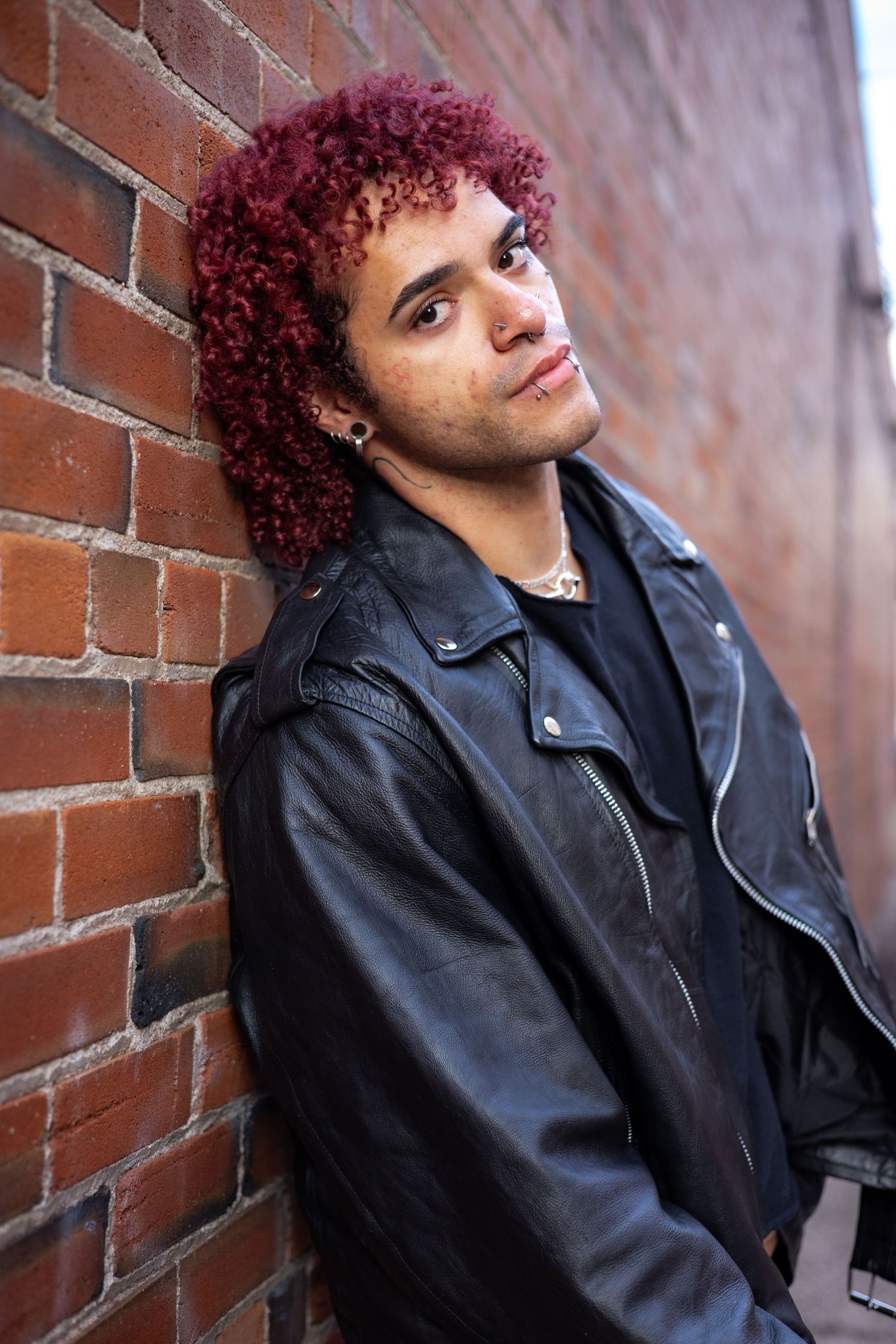 Person with vibrant red curly hair poses against brick wall wearing leather jacket.