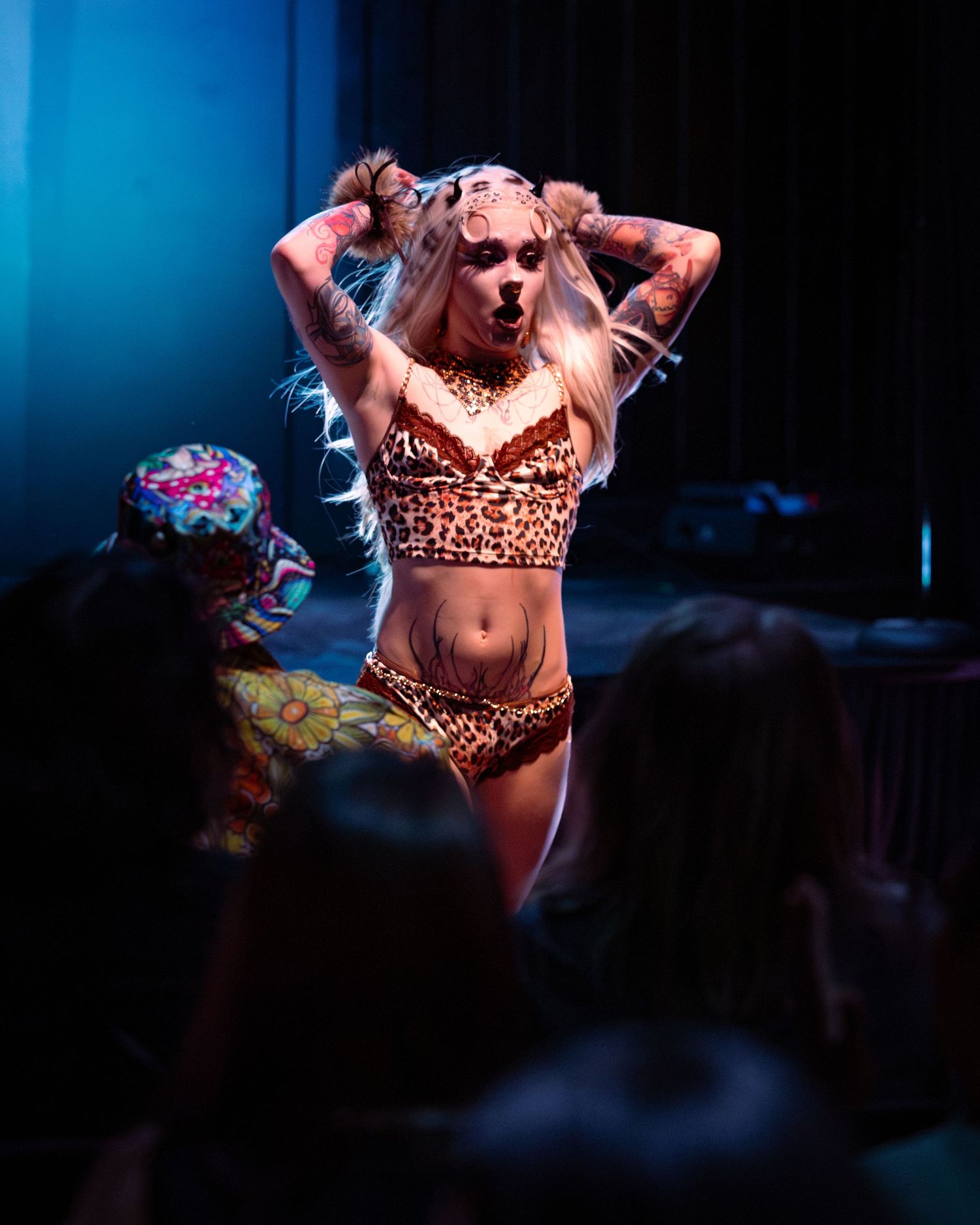 Tattooed blonde performer in leopard print outfit dancing before a crowd under blue stage lighting.