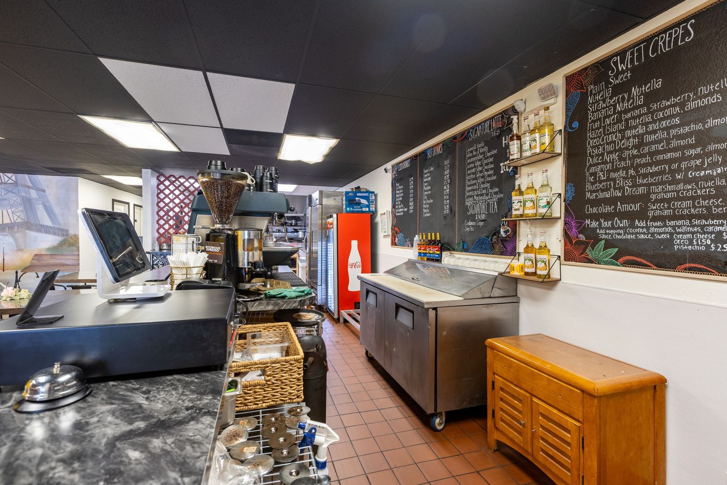 Interior view of a cozy cafe counter with POS system, espresso machine, chalkboard menu, and display cases on tile floors.