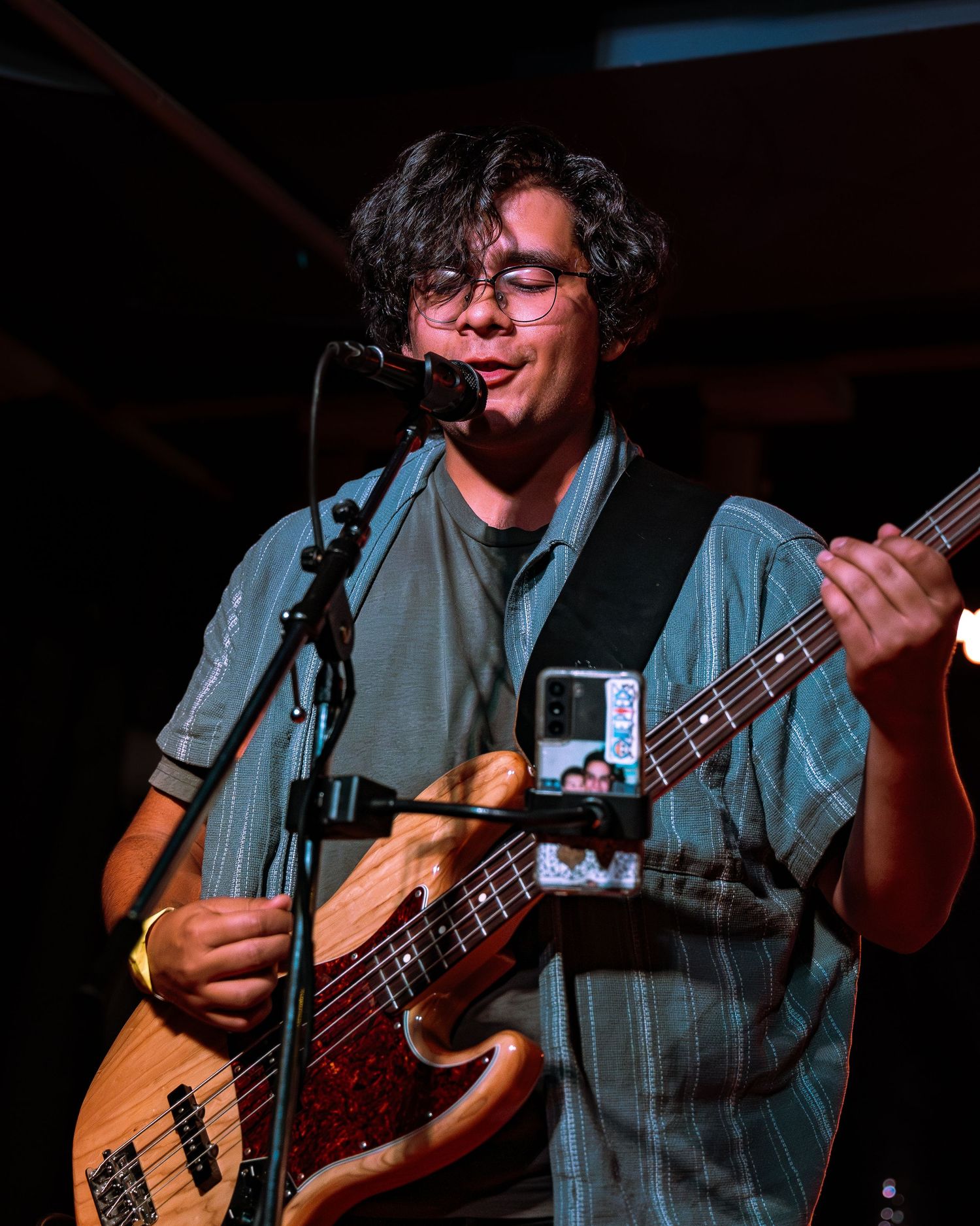 Smiling bassist with glasses sings into microphone while playing a wood-finish bass guitar at live show.