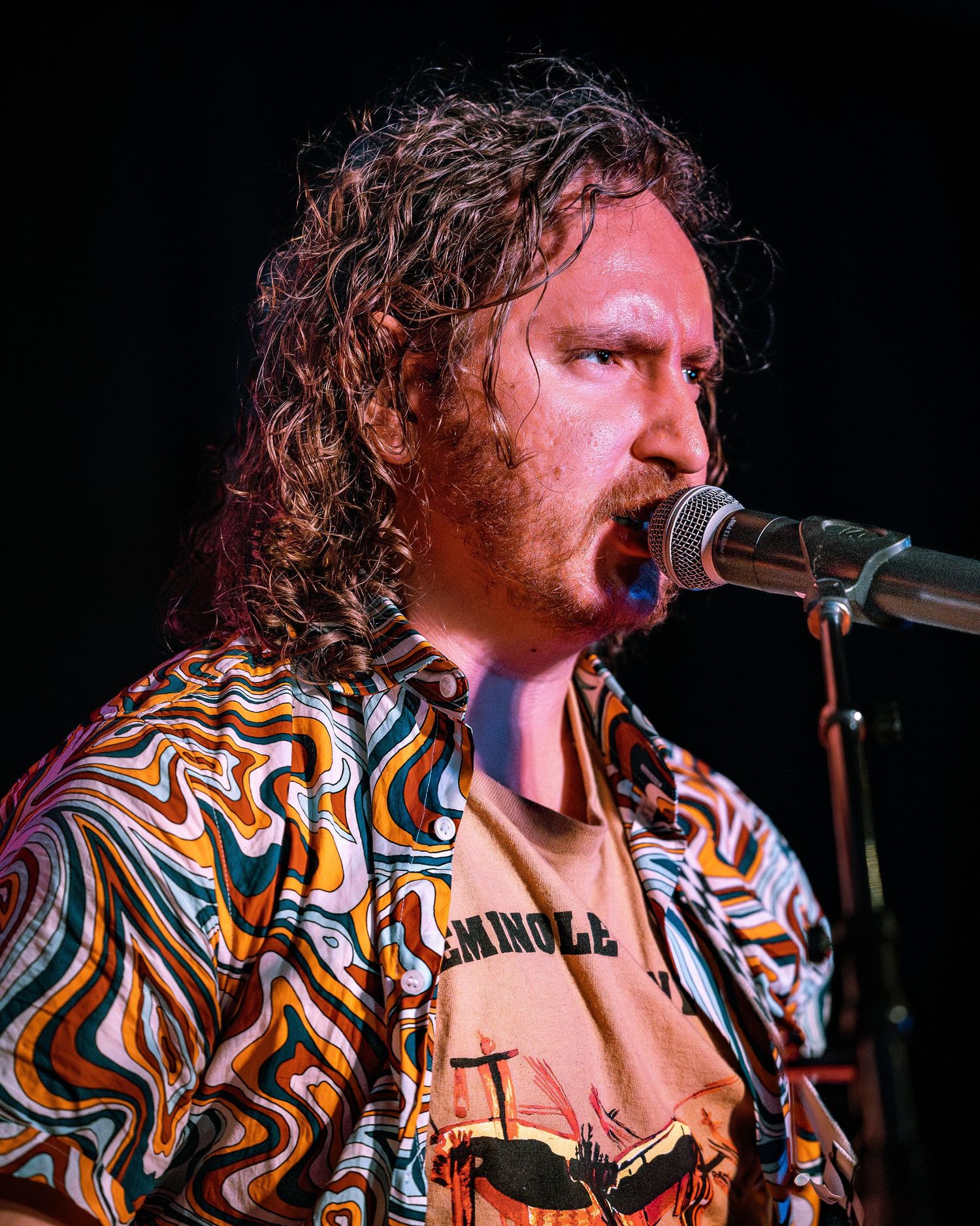 Curly-haired male singer performs passionately in colorful swirl-patterned shirt on stage.