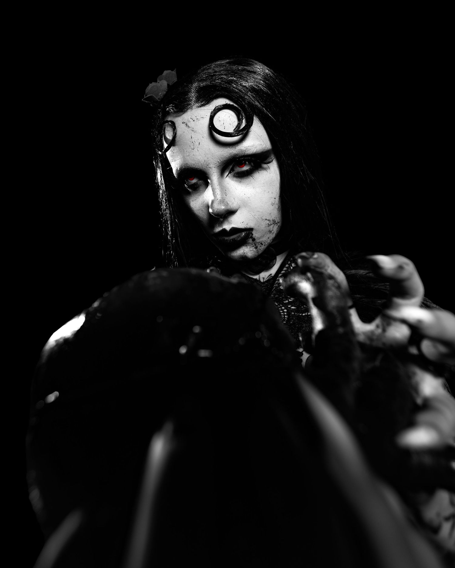 Black and white gothic portrait with dramatic dark makeup, spiral forehead symbol, and red eyes in moody lighting.