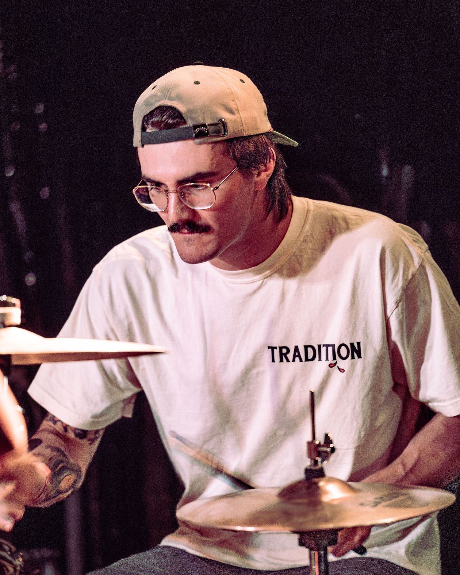 Tattooed drummer with mustache, glasses and snapback cap playing cymbals on stage.
