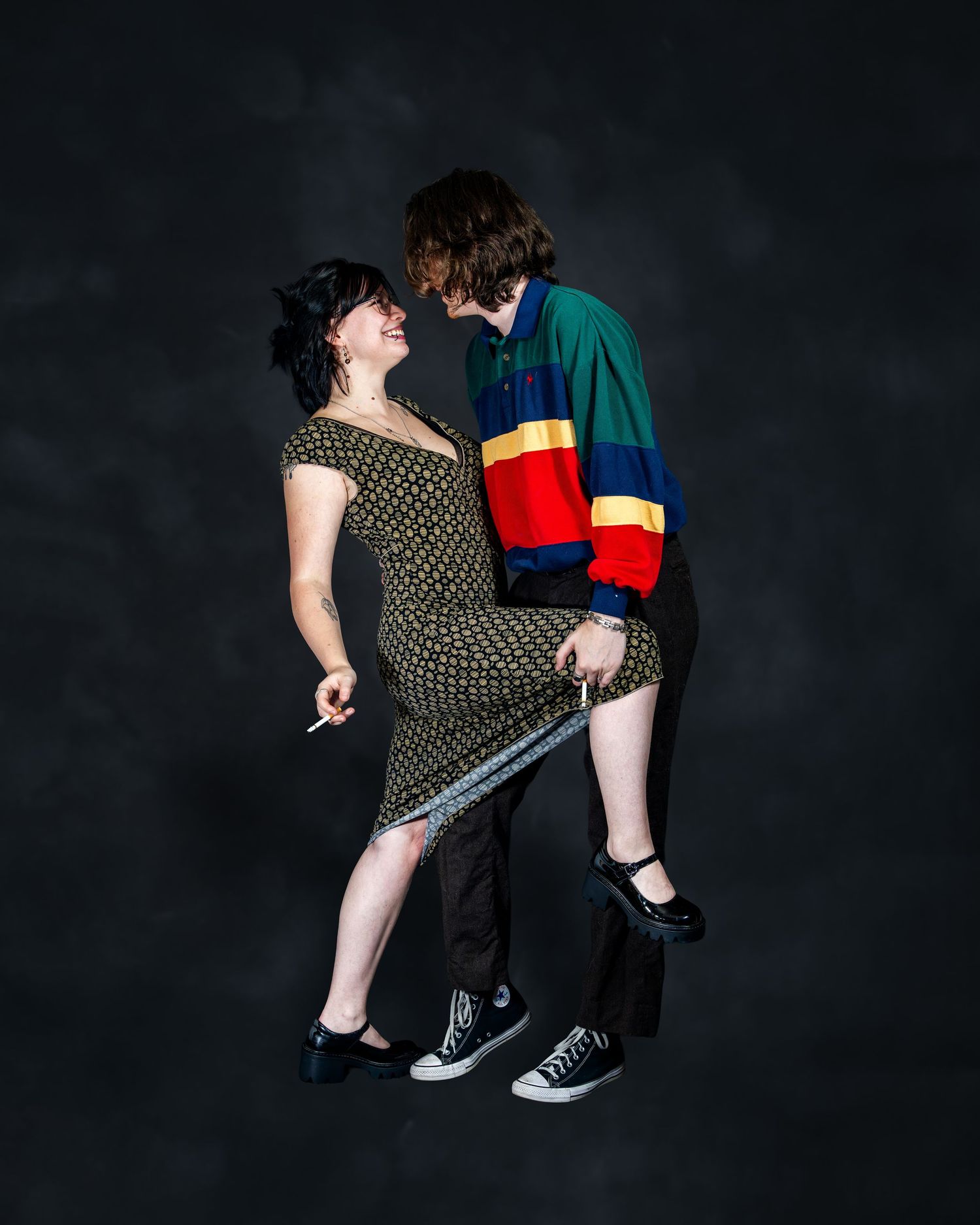 Two young people in colorful retro outfits lean in to kiss against a dark studio background.