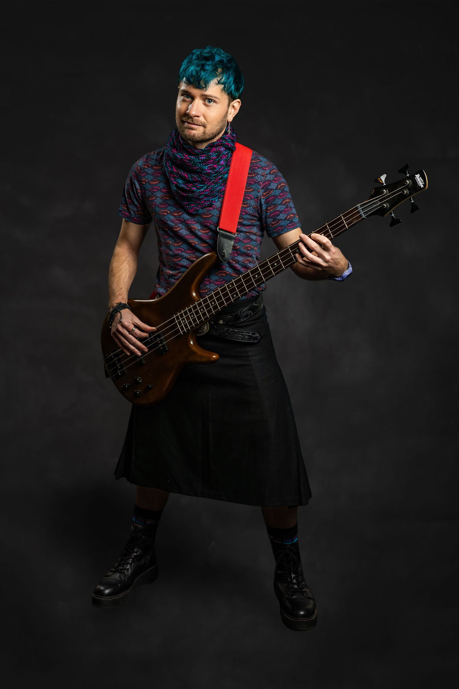 Man with teal hair playing bass guitar wearing kilt and red strap in dark studio.
