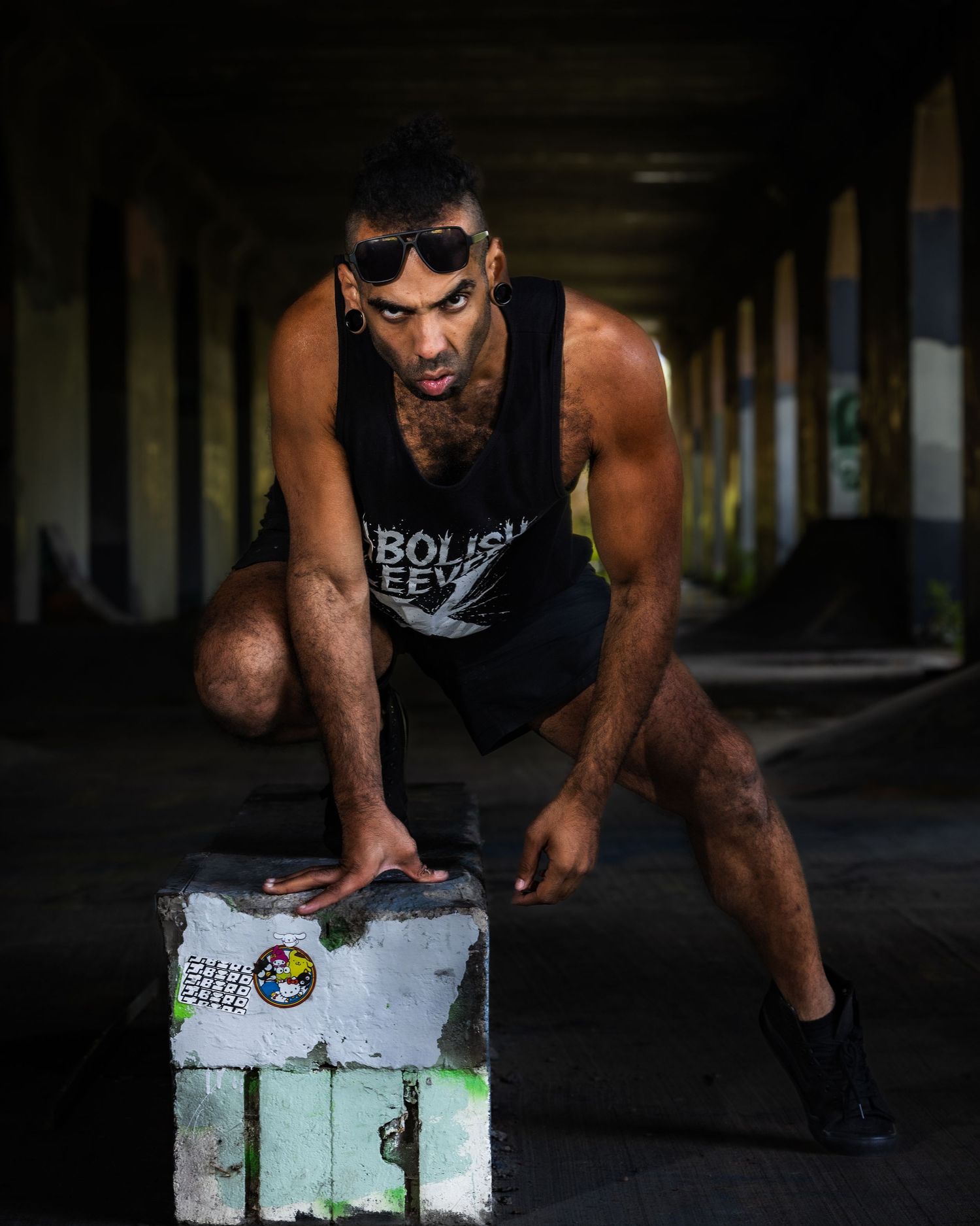 Athletic person in a dark tank top and sunglasses crouches on a painted concrete block in a dimly lit urban setting.