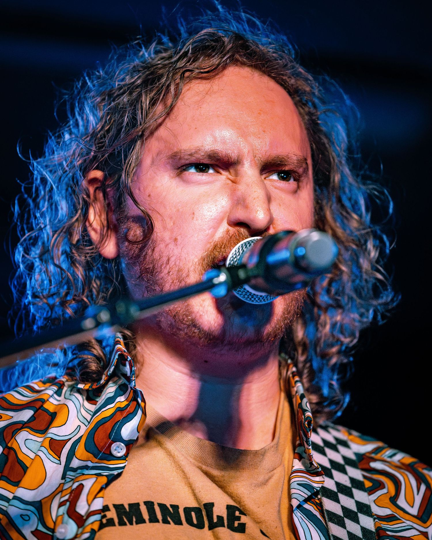 Curly-haired male singer performing live on stage, wearing a colorful Seminole patterned shirt while singing into a microphone.