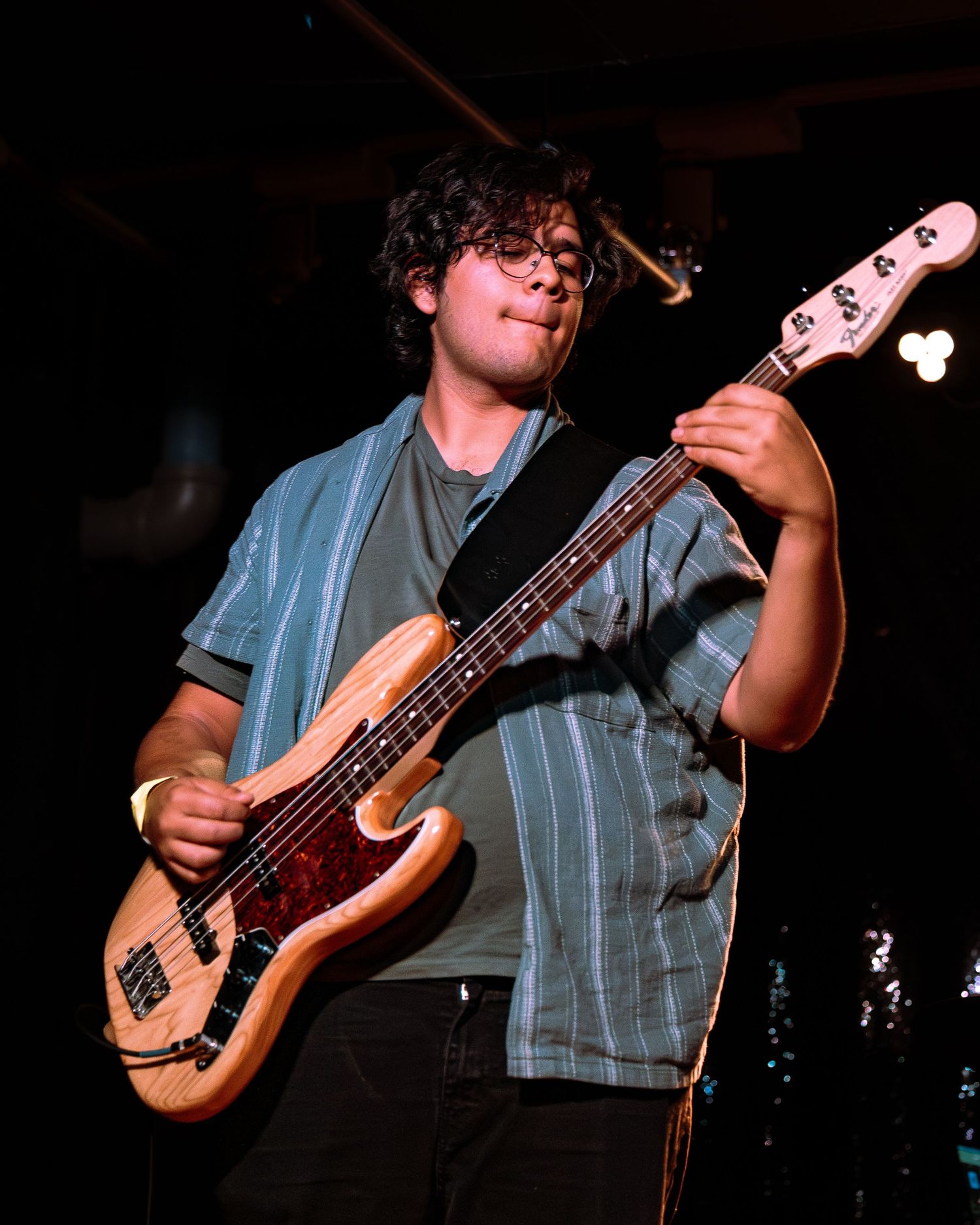 Young musician playing a wooden bass guitar on stage under bright lights wearing a blue striped shirt.