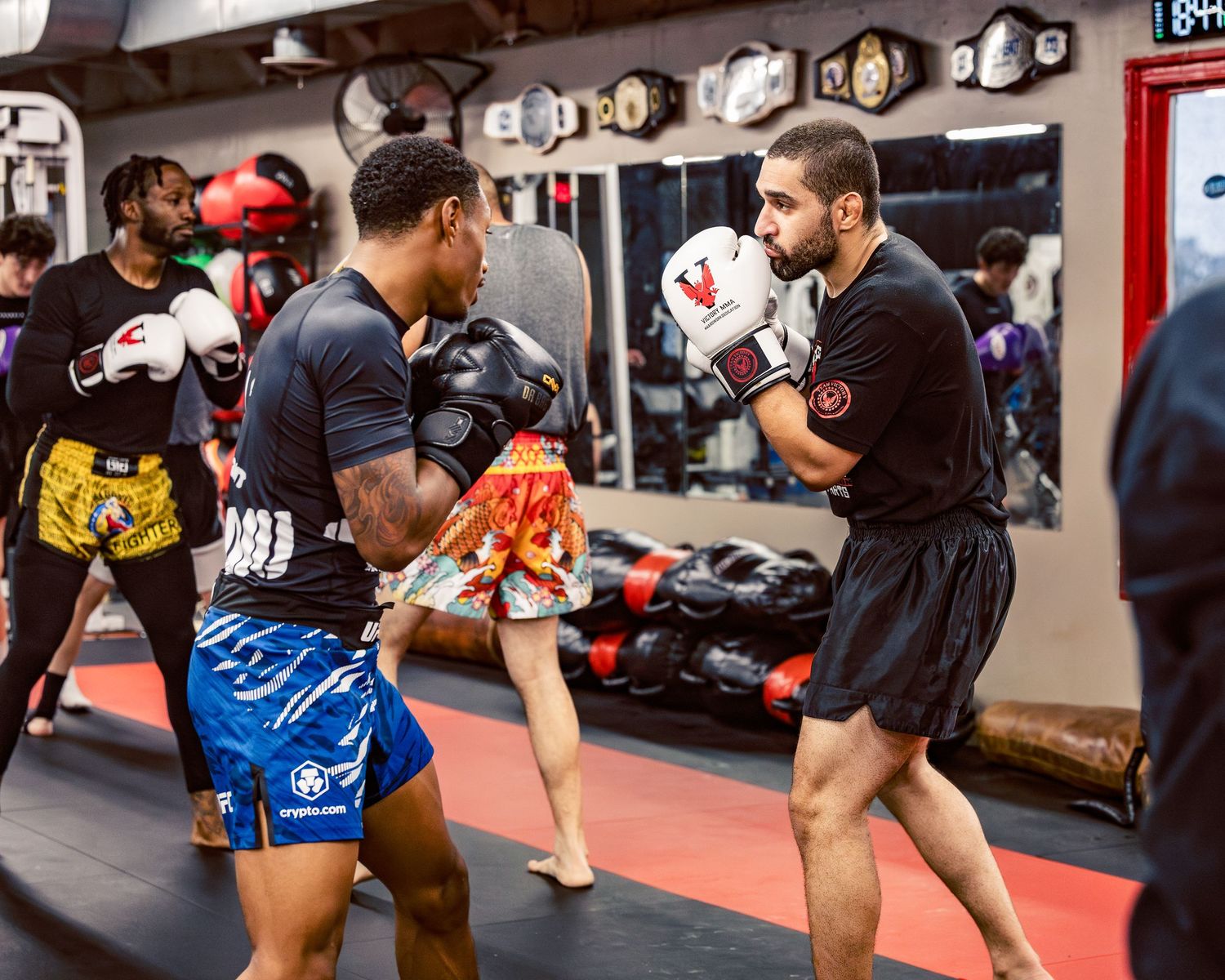 Athletes performing martial arts drills on red mats with fight equipment visible in the background.