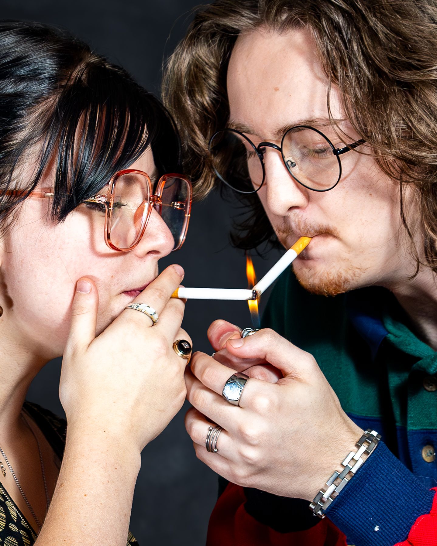 Two people with glasses lighting cigarettes together, sharing a flame from a lighter in close proximity.