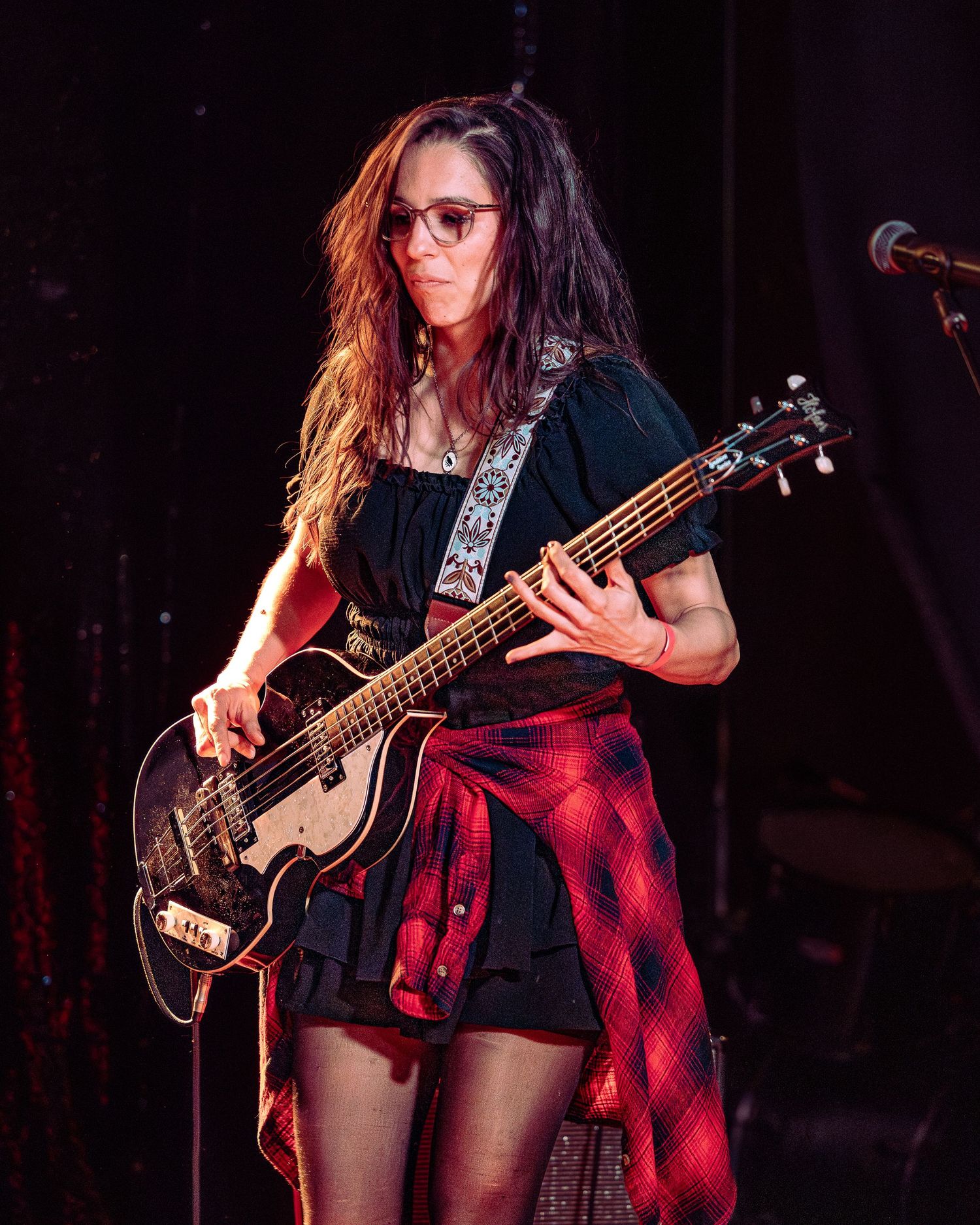 Woman with long hair wearing glasses playing black bass guitar with red flannel tied at waist.
