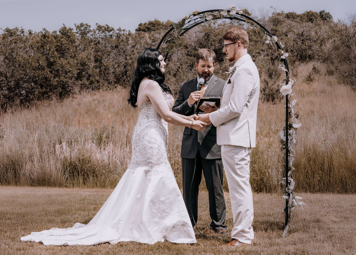 A couple shares their wedding ceremony in a rustic outdoor setting with brown grass and trees in the background.