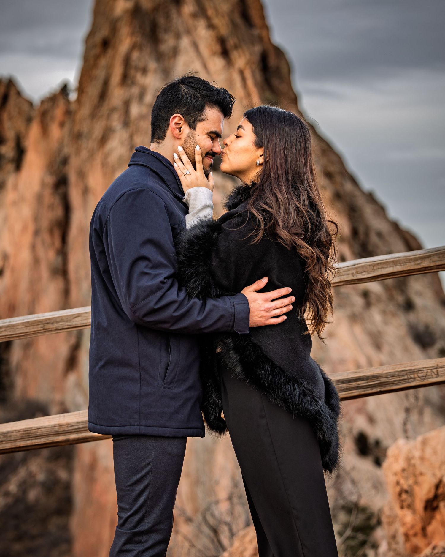 A romantic couple embraces and kisses on a wooden walkway with dramatic red rock formations in the background.
