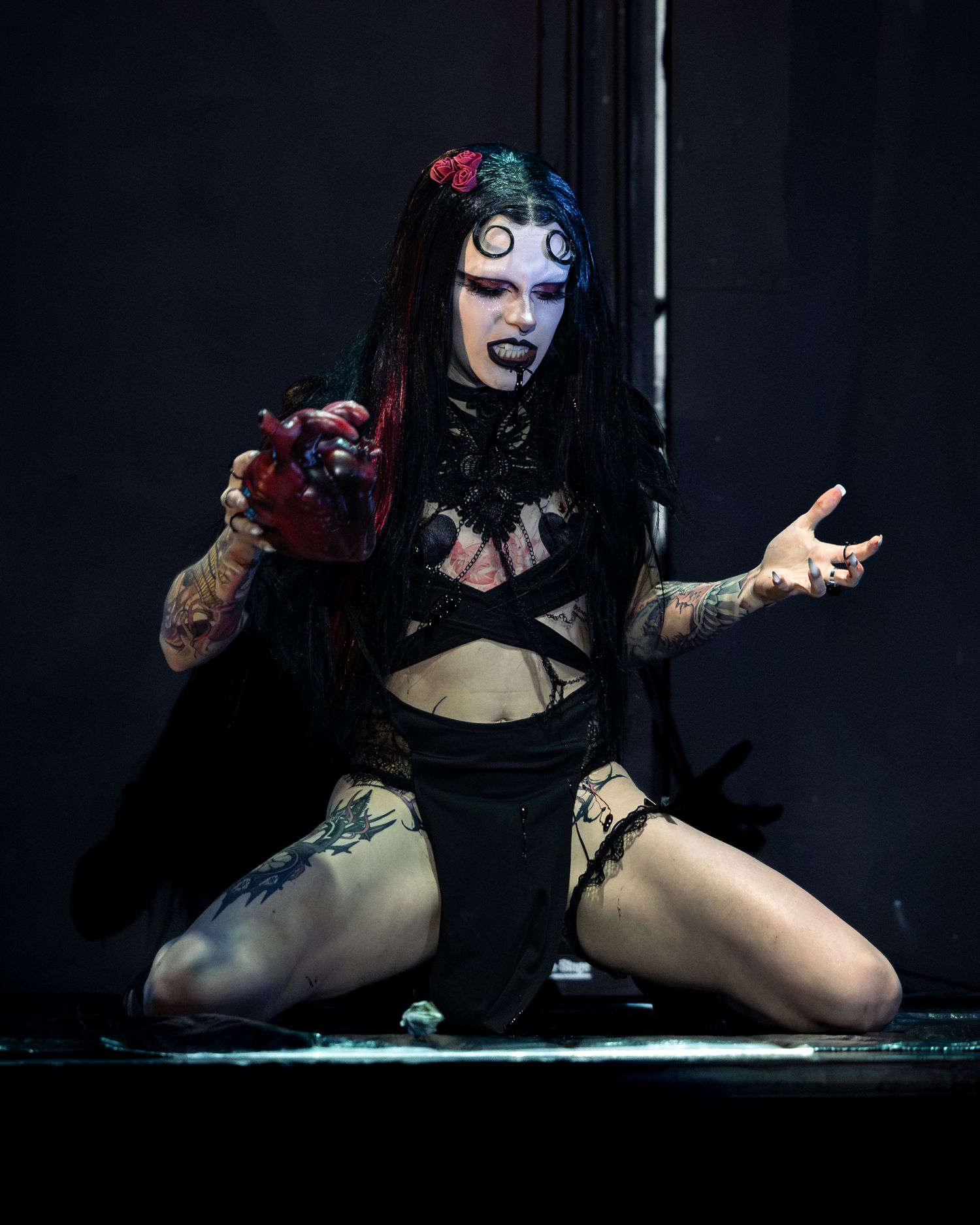 Tattooed gothic performer kneels on stage holding bloody heart with dramatic black and red makeup.