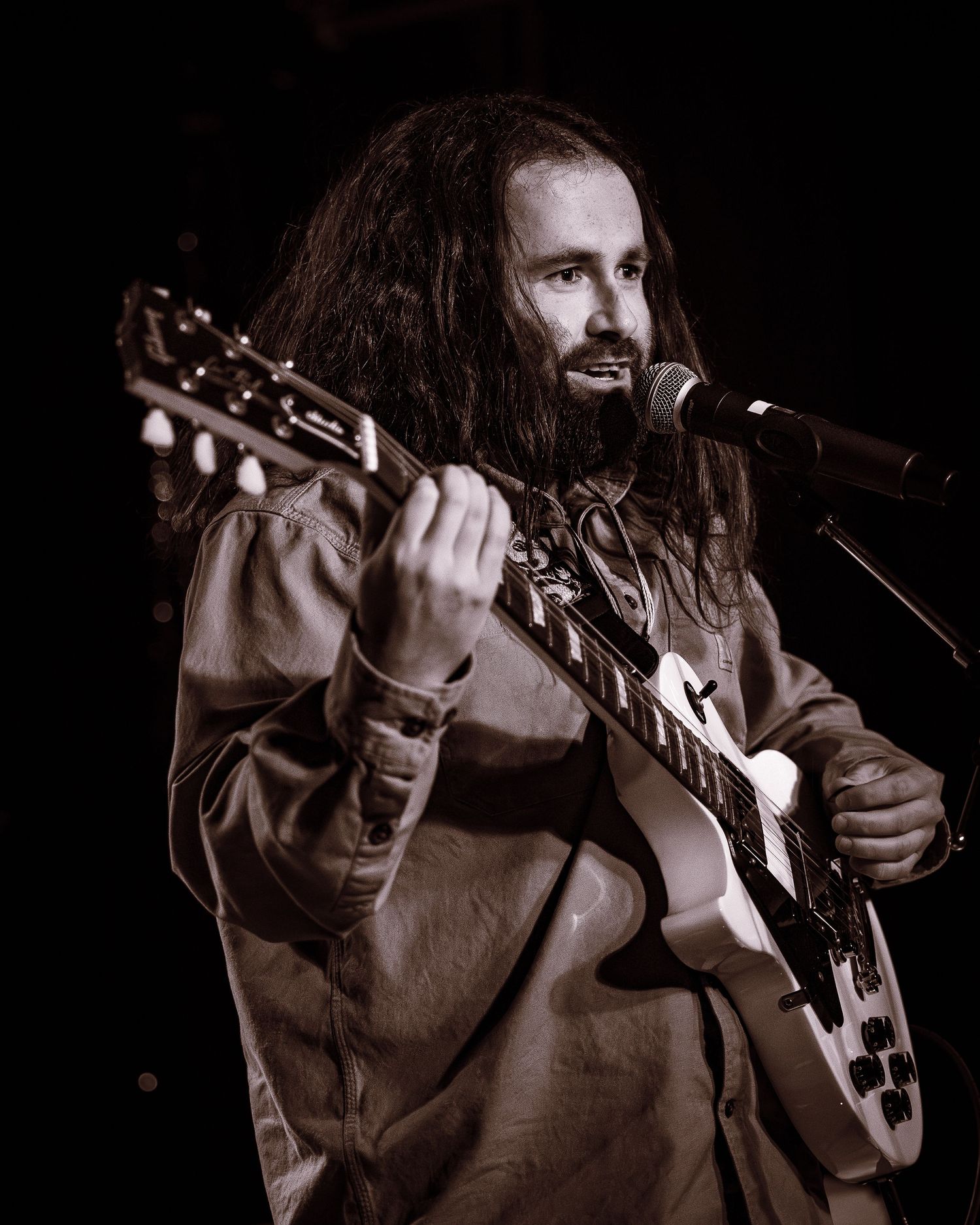Long-haired bearded guitarist singing into a microphone while playing an electric guitar.