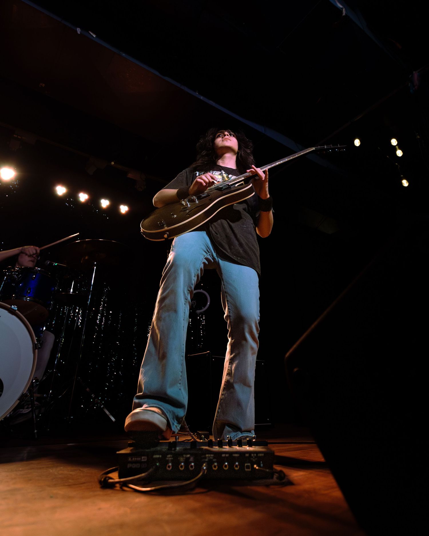 Guitarist performs on stage in blue flared pants, playing a dark electric guitar under bright concert lights.