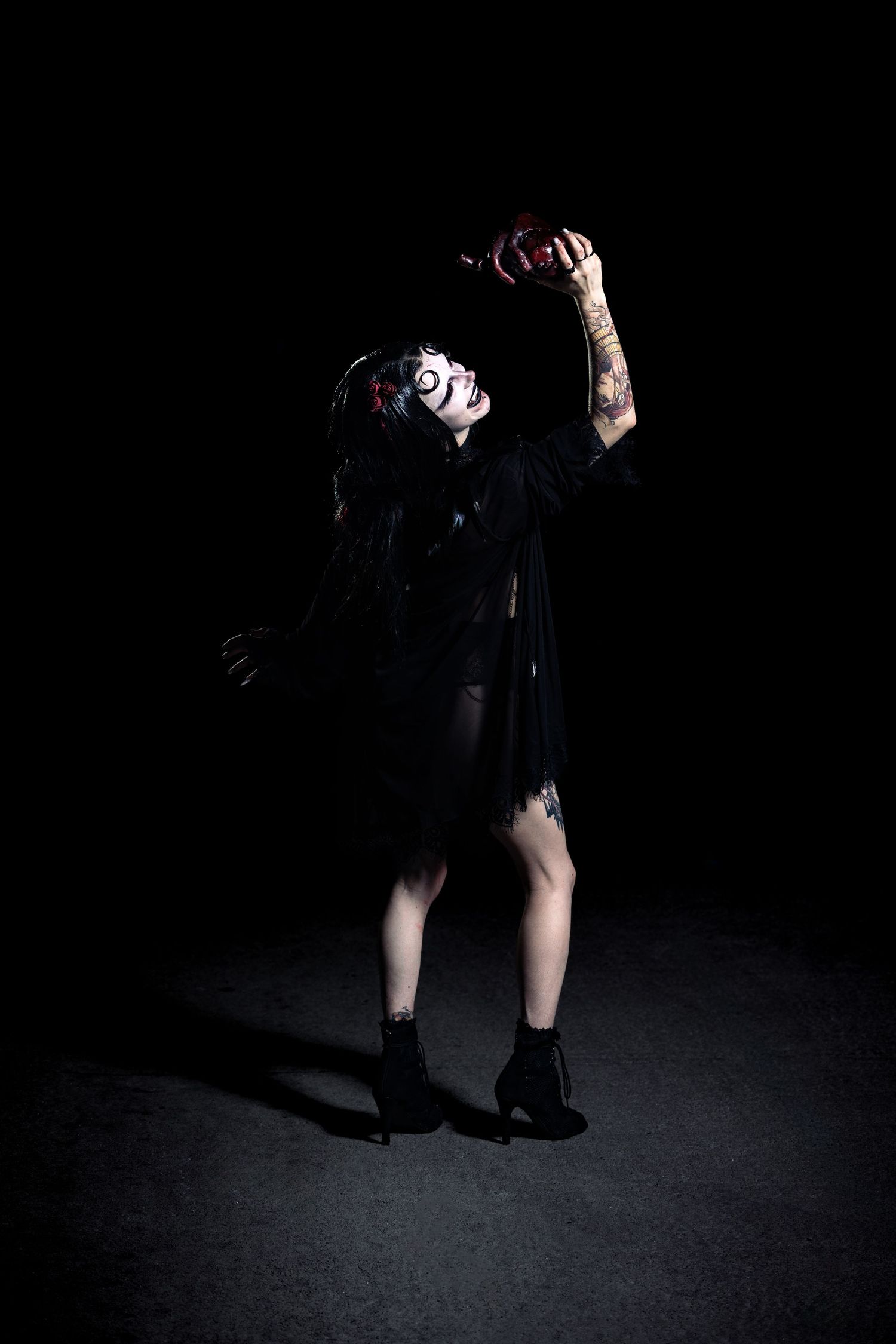 Tattooed woman in torn black dress with zombie face paint holding bloody prop aloft in dark dramatic scene.