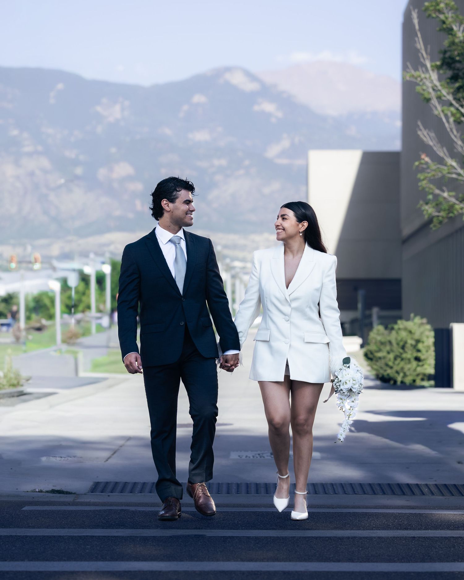 Couple in wedding attire holding hands walking outdoors with mountain backdrop, bride in white blazer dress, groom in dark suit.