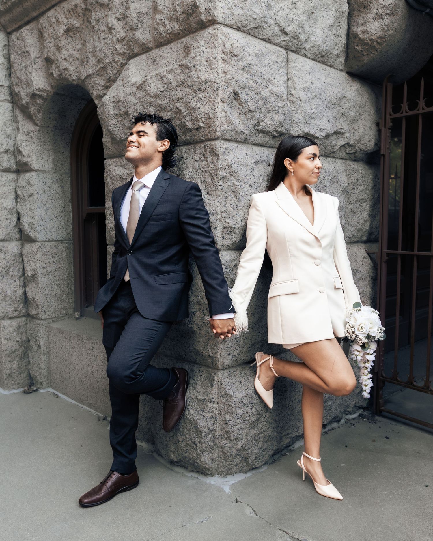 Couple in wedding attire holding hands against stone building, groom in navy suit, bride in white blazer dress with bouquet.
