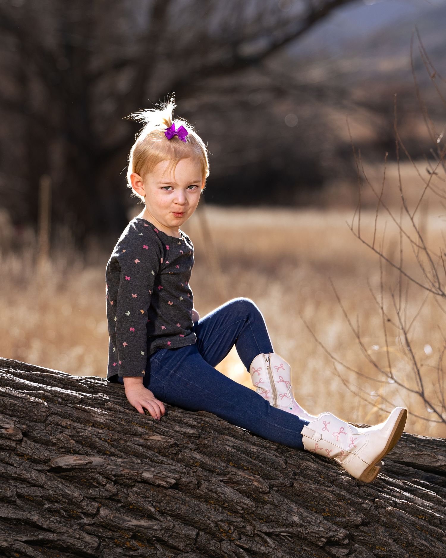 Cute blonde girl with purple hair bow sitting on a fallen log in a rustic outdoor autumn setting.