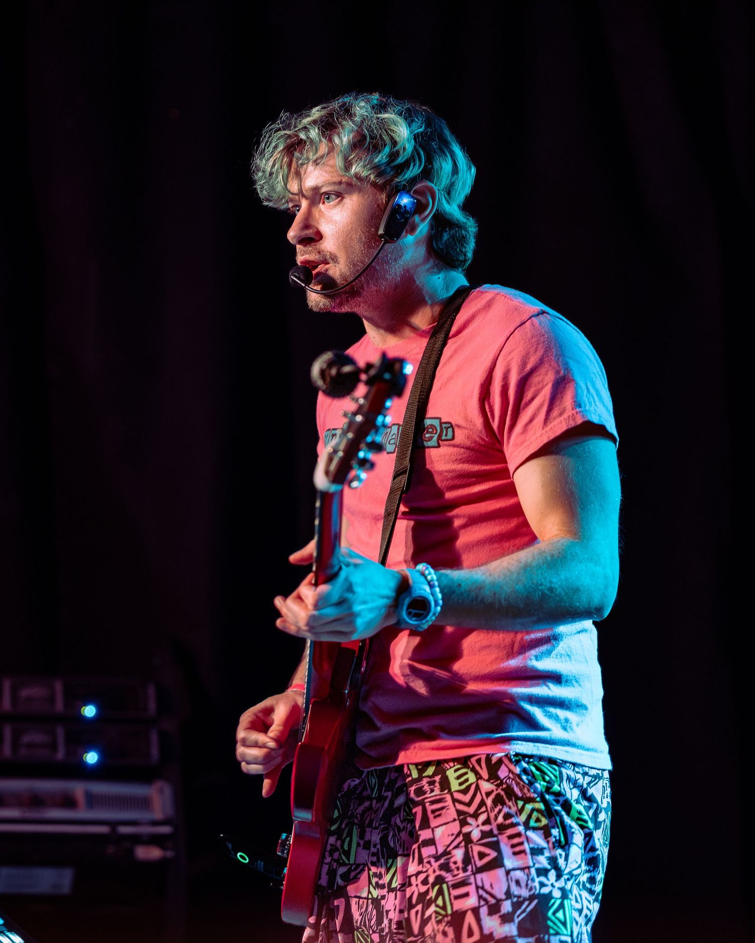 Musician playing red electric guitar on stage wearing pink t-shirt and colorful shorts with headset microphone.