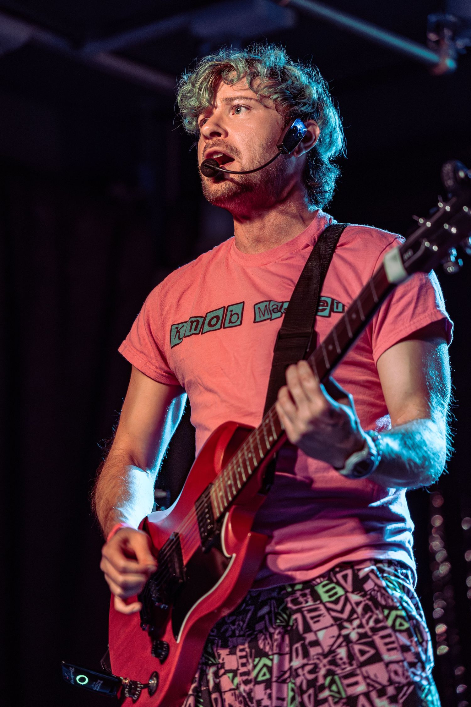 Male guitarist performing live on stage in pink Knob Creek t-shirt and patterned shorts with headset mic.
