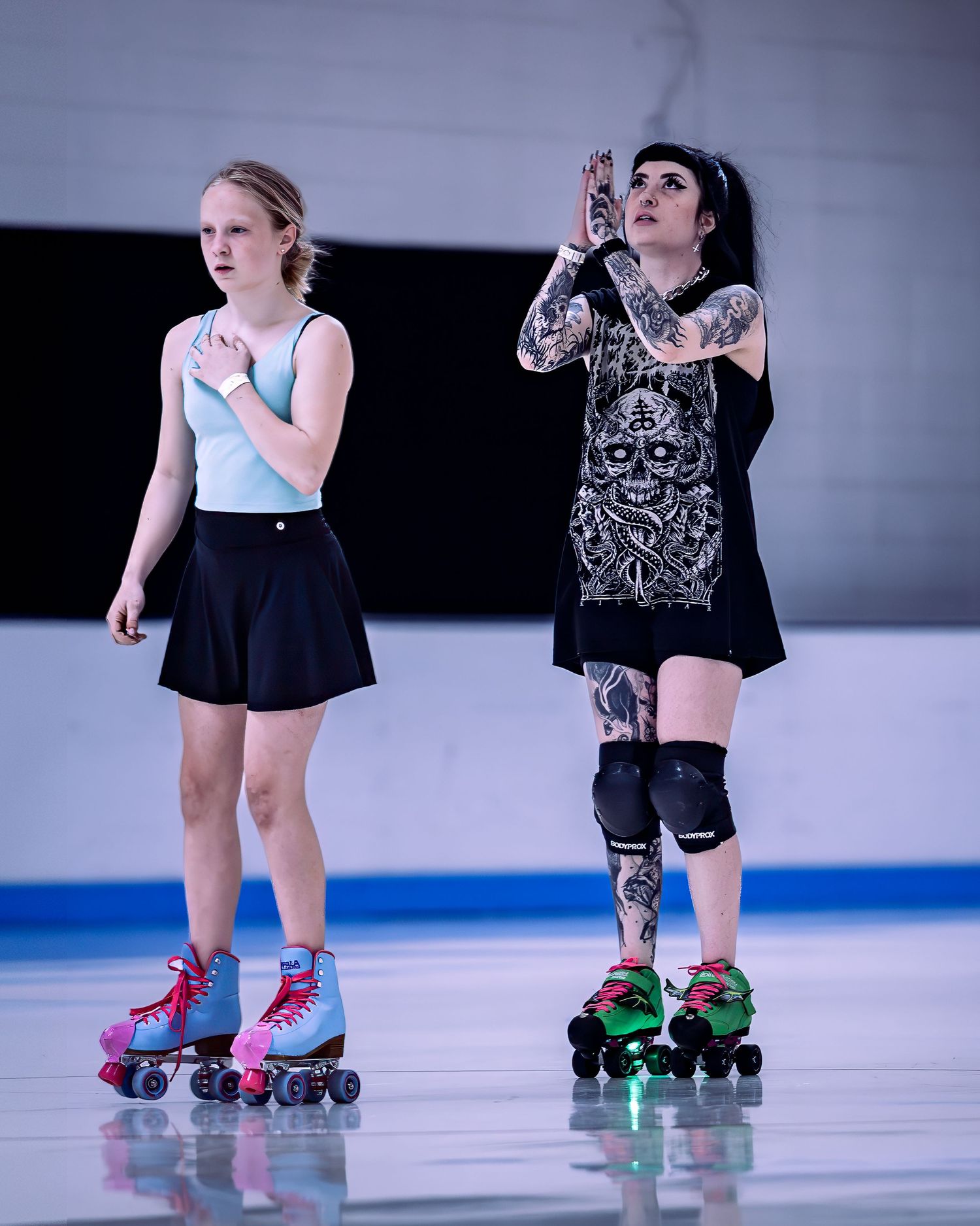 Two roller derby skaters in black outfits with colorful quad skates practice moves on an indoor rink.