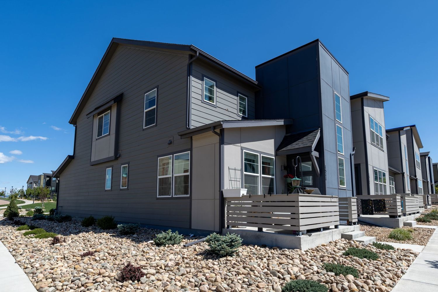 Modern two-story townhome with gray and dark exterior, landscaped rock yard, and covered porch under a bright blue sky.