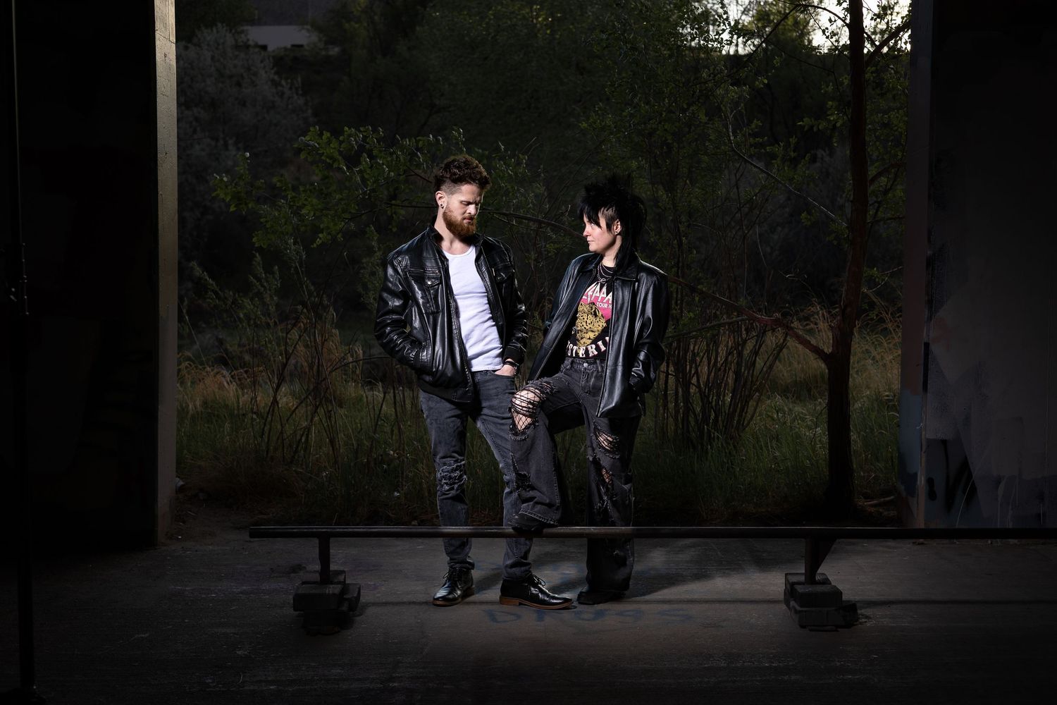 Two men in black leather jackets stand facing each other outdoors at night in a dramatic, edgy pose.