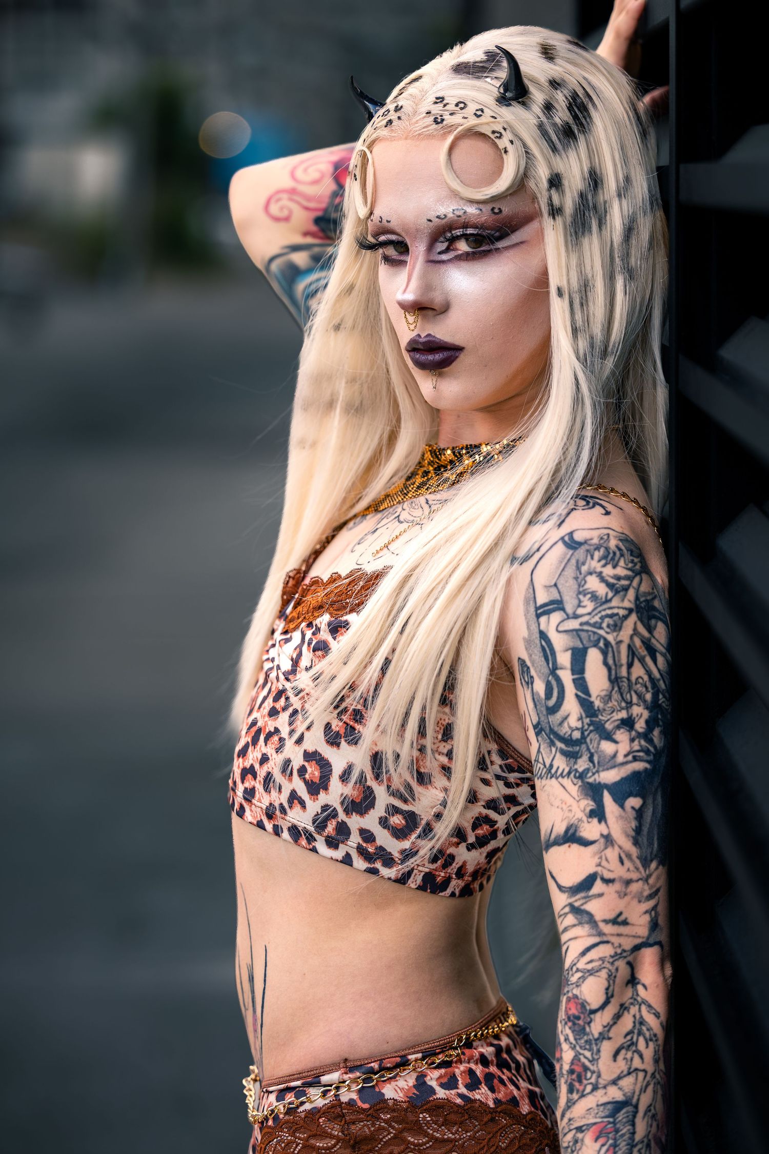 Tattooed blonde woman in leopard crop top with dramatic face paint and horns leaning against dark wall.