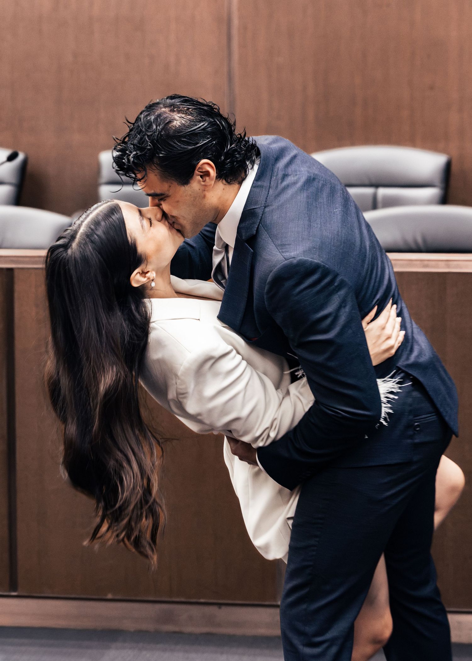 Couple sharing a romantic kiss in a courtroom, dressed in formal attire, with wooden paneling in the background.