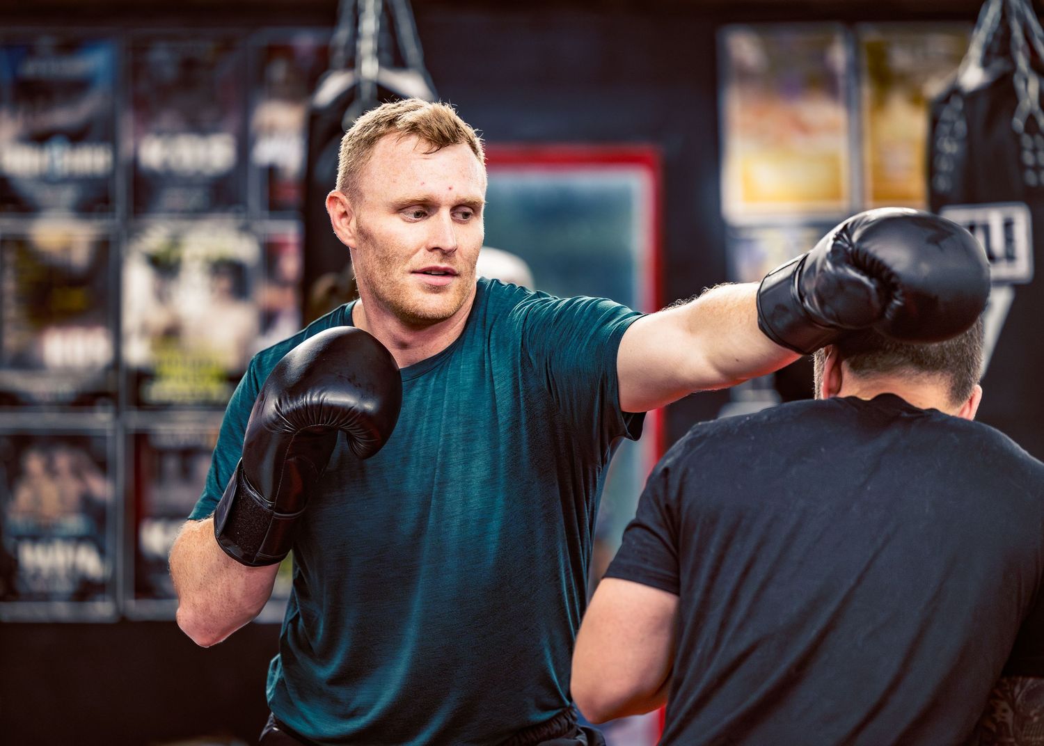 Two athletes practice boxing techniques in a gym with fluorescent lighting overhead.