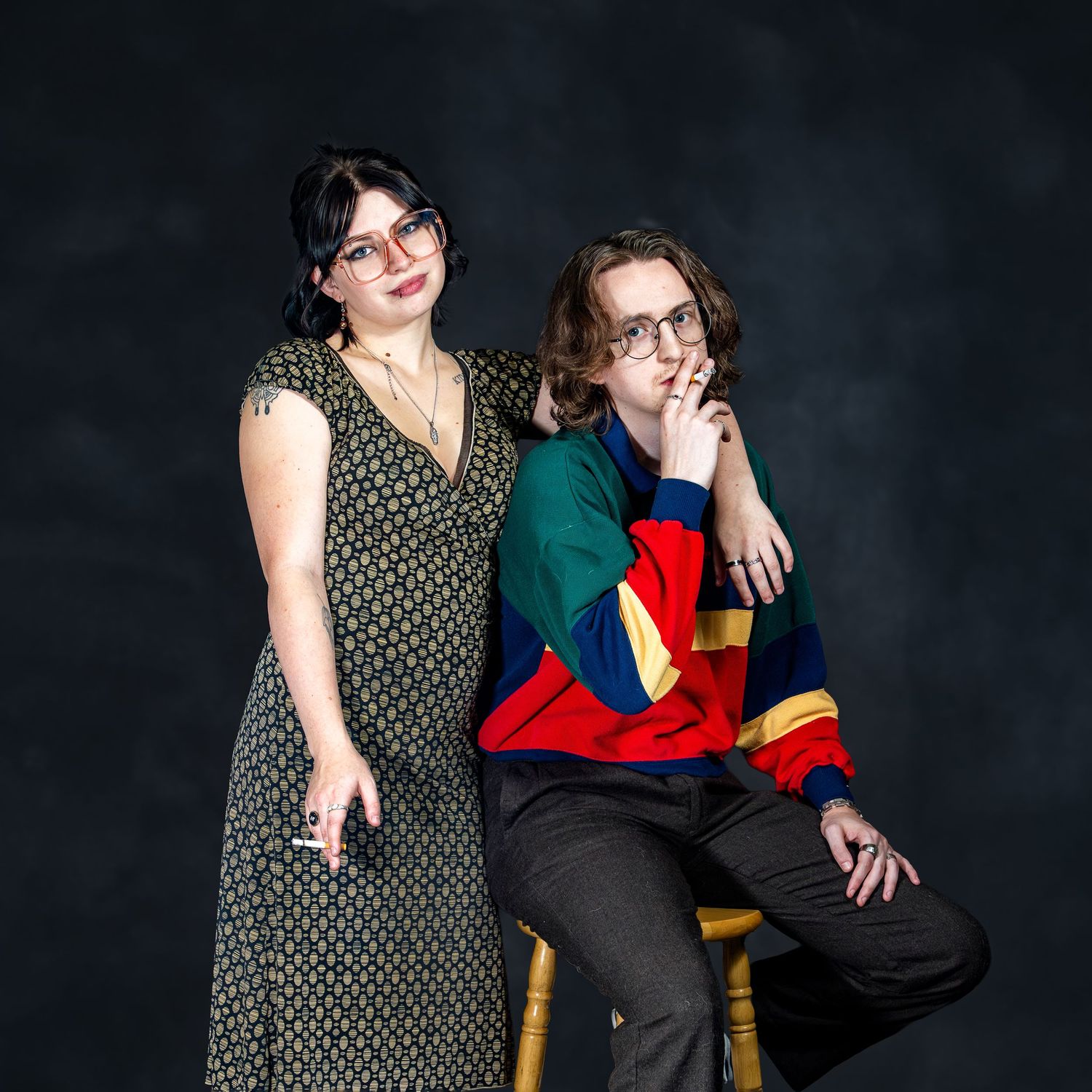 A woman in floral dress and man in colorful striped sweater pose in a dark studio setting.